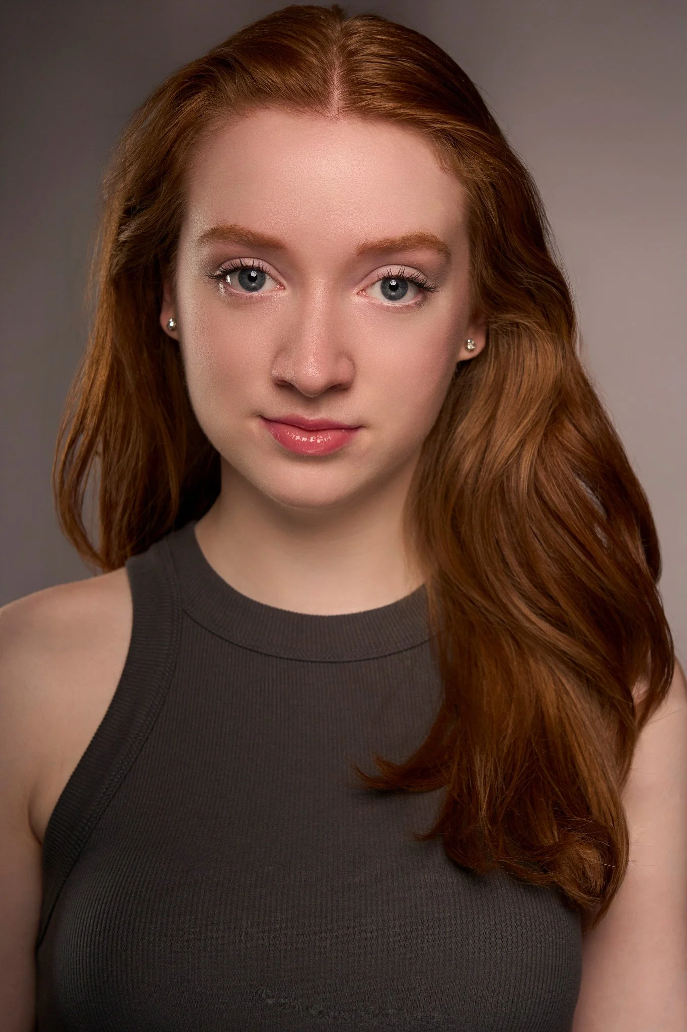 Close-up portrait of a young woman with curly red hair, blue eyes, and fair skin, wearing a black sleeveless top and small earrings, against a gray background.