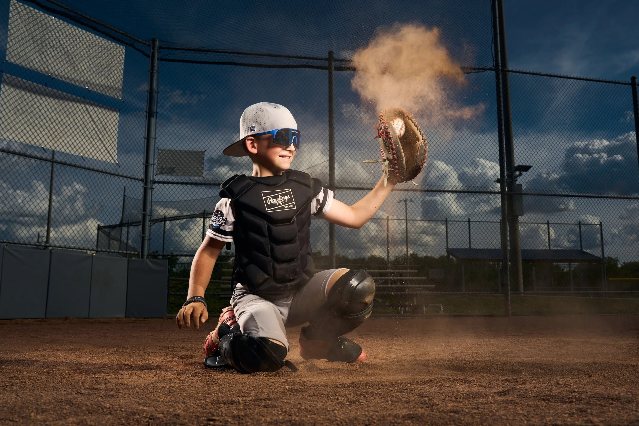 A young baseball player kneeling on the dirt in a baseball field, with a catcher's mask hanging from his hand, wearing a catcher's chest protector, a cap worn backwards, sunglasses, and showing a smile. The boy has just caught a baseball, which is saved in a glove, and a puff of dust is visible in the air behind the ball. The background features a chain-link fence and a partly cloudy sky.