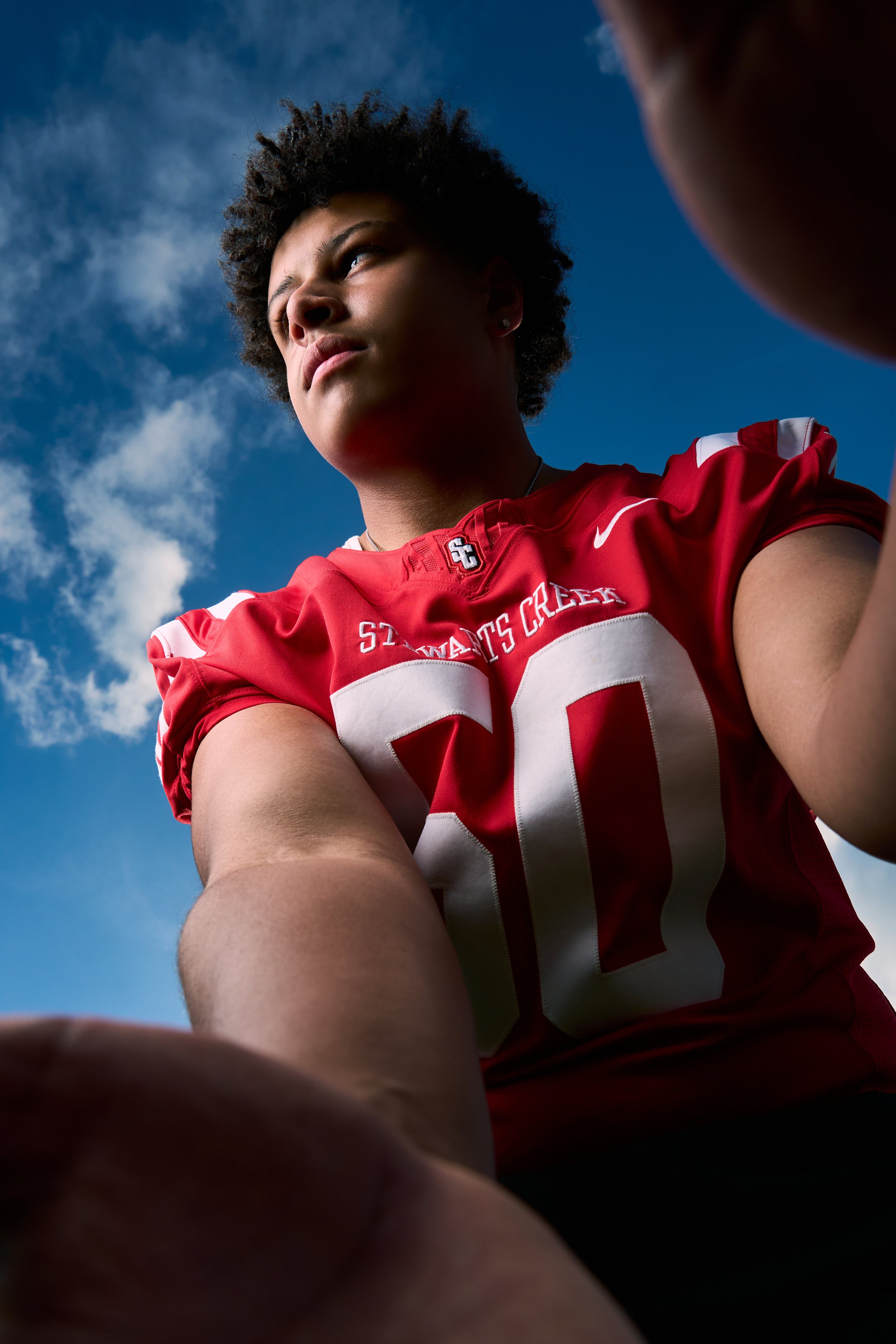 A female athlete in a red sports jersey with white stripes, looking upwards against a blue sky with clouds, taken from a low angle.