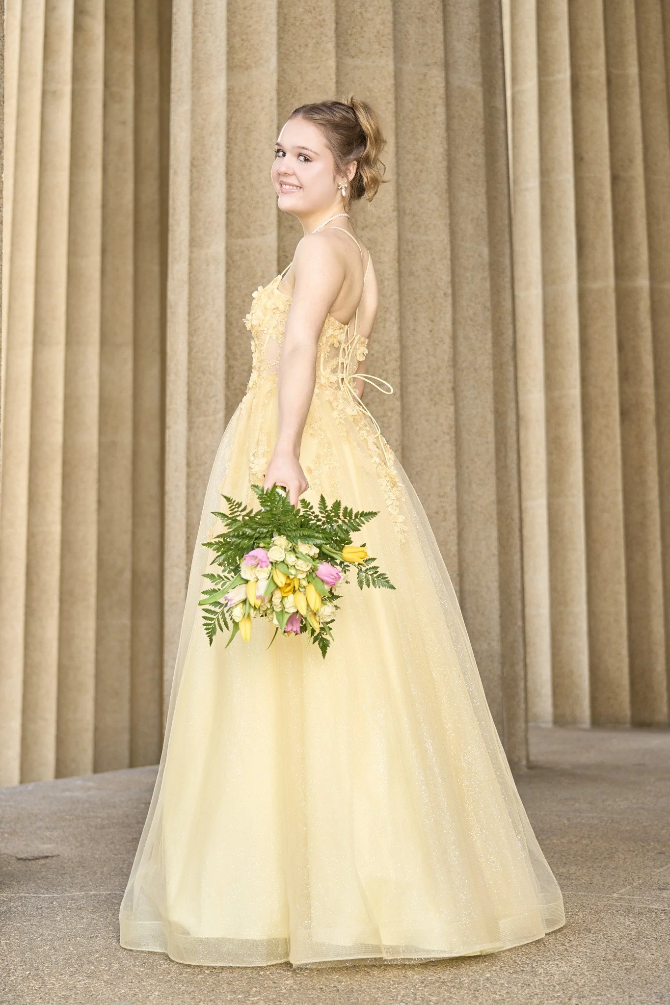 Young woman in a yellow gown holding a bouquet of pink, yellow, and white flowers stands in front of beige curtains, smiling partially toward the camera.