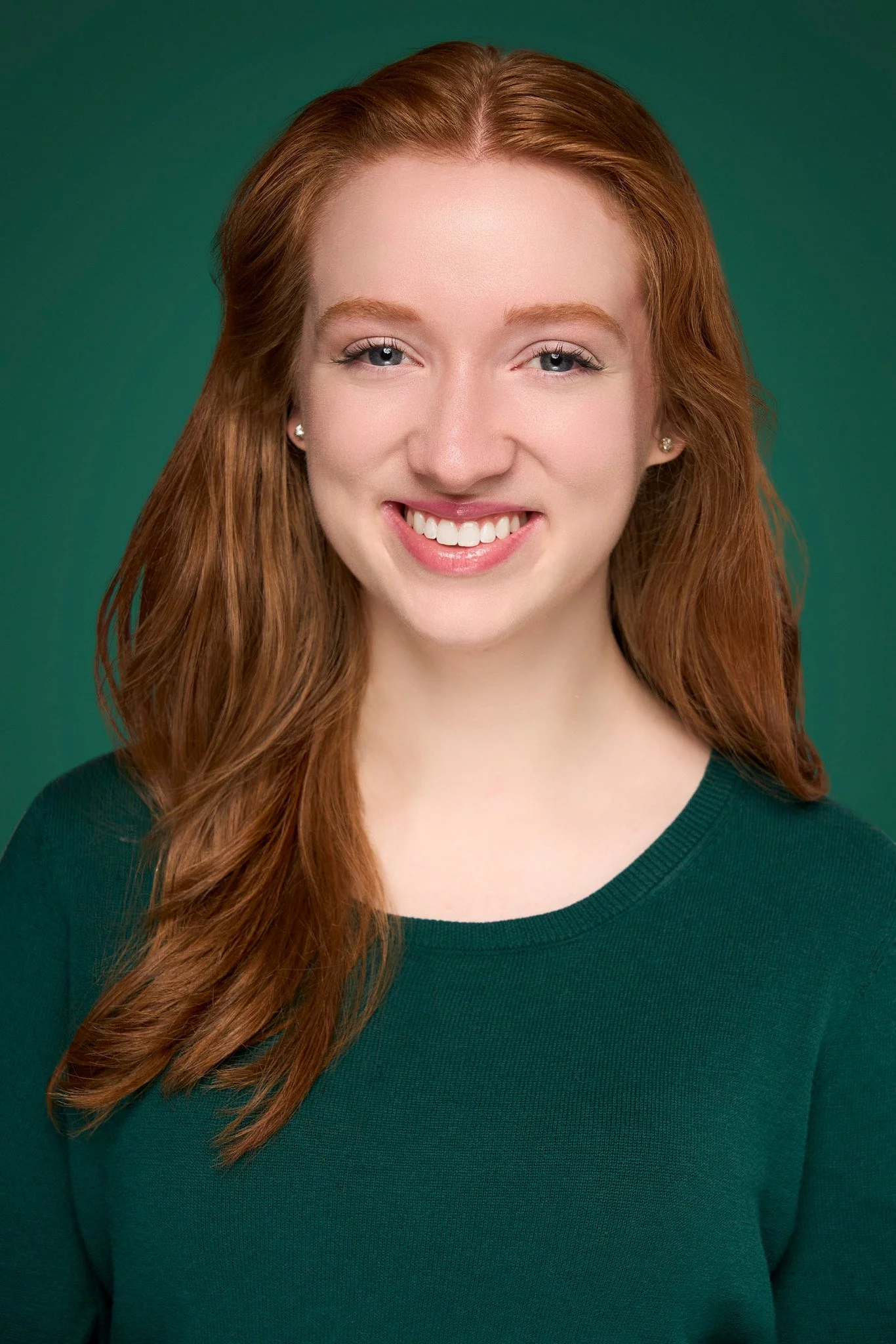 Close-up of a woman with long red hair, smiling, wearing a dark green top, against a matching green background.