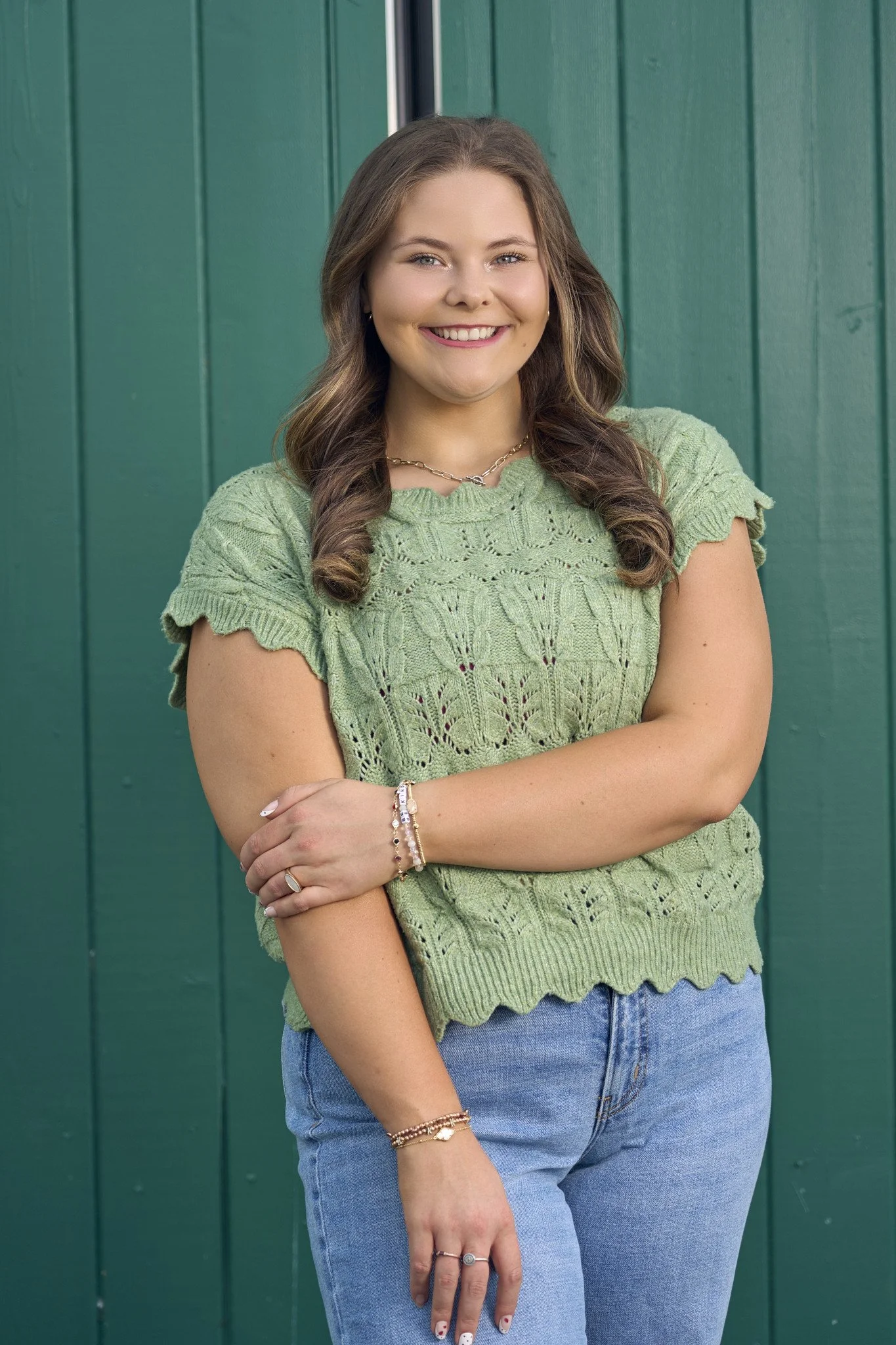 A young woman with light brown hair and a bright smile is standing in front of a green wooden wall. She is wearing a light green crocheted top and blue jeans. She has jewelry including rings, bracelets, and a necklace.