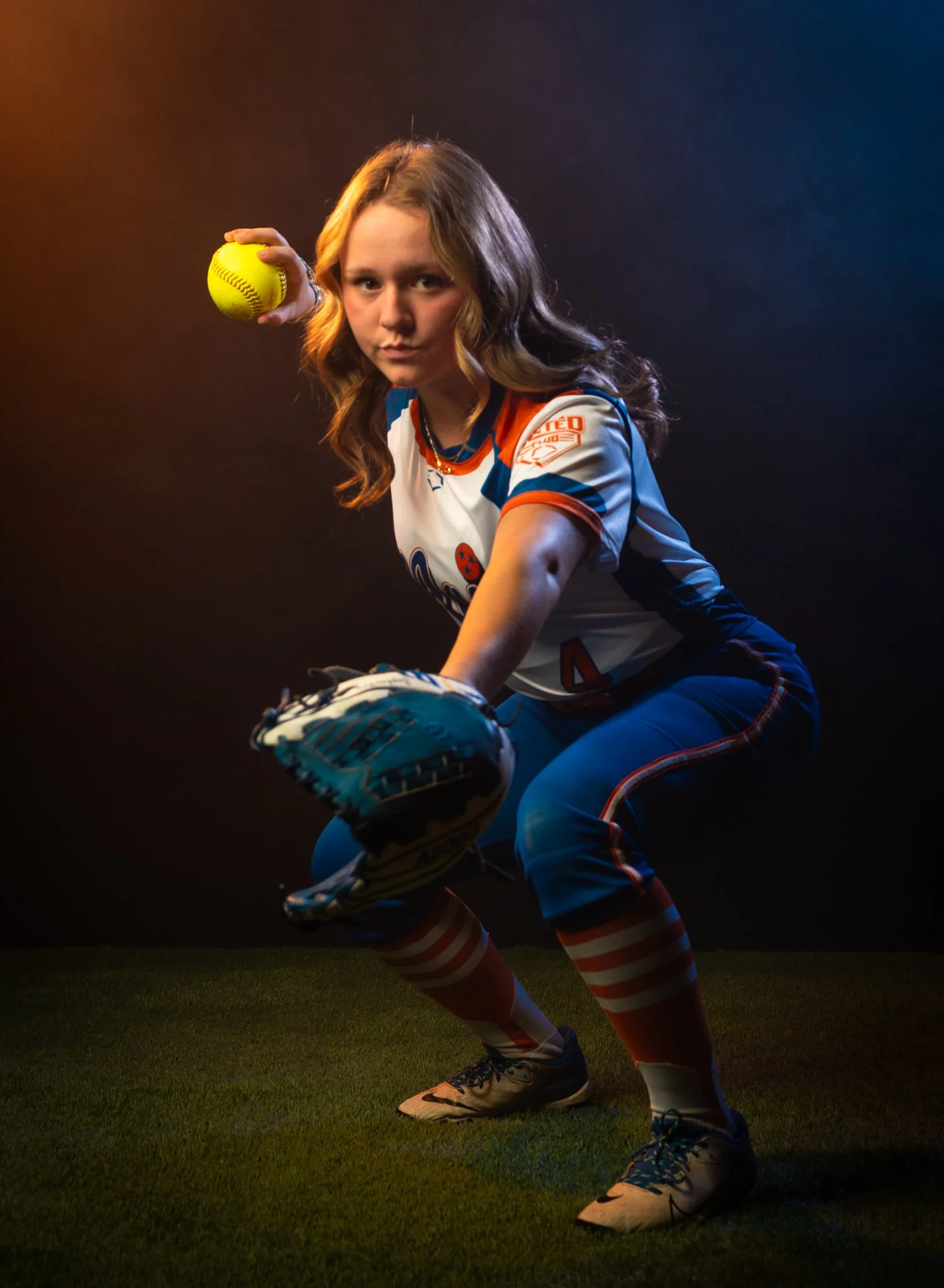 A young female softball player wearing a blue, white, and orange uniform with striped socks is in a crouched pose, holding a softball in her right hand and wearing a glove on her left hand. The background is dark with a gradient of orange and blue li