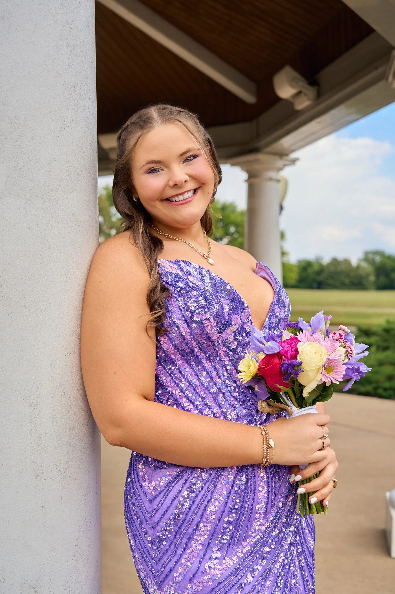 A young woman in a purple, sequined dress holding a bouquet of colorful flowers, smiling, standing outdoors near a white pillar with a wooden pavilion and lush green trees in the background.