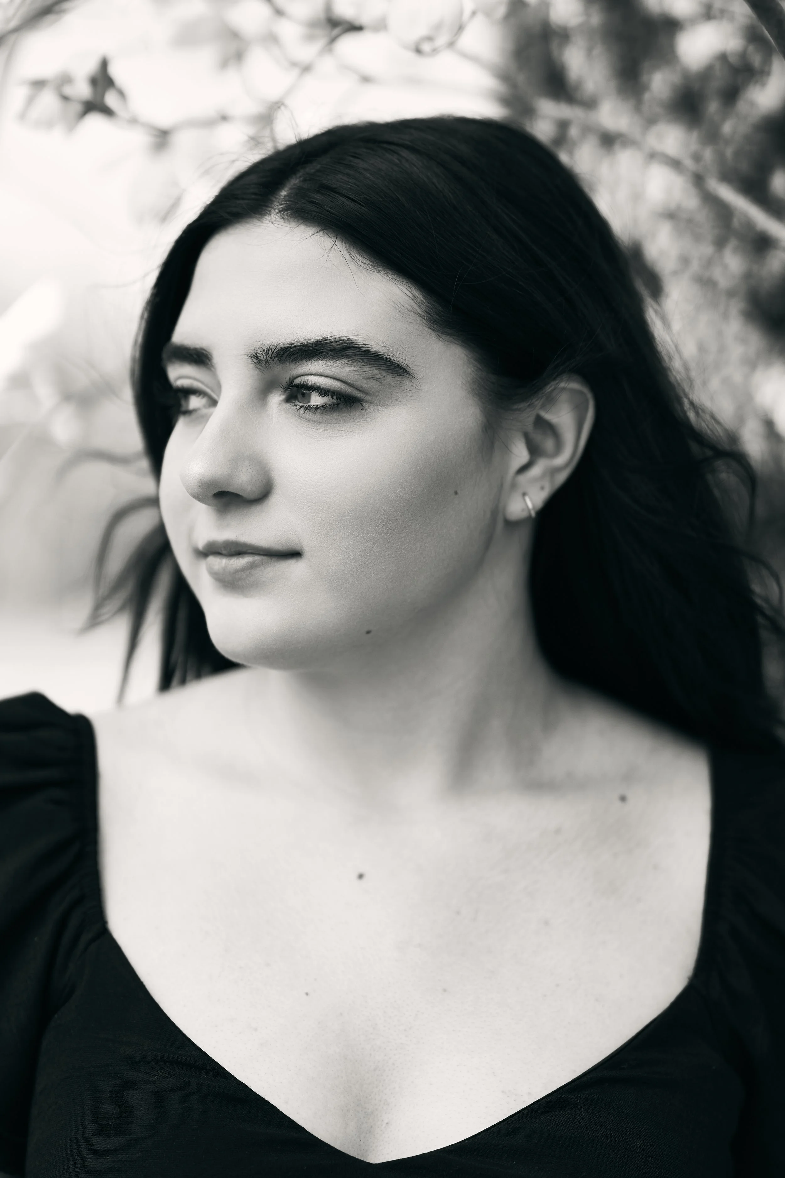 A black and white photo of a woman with dark hair, looking to her left, dressed in black, with a slightly smiling expression, in front of blurred natural background.
