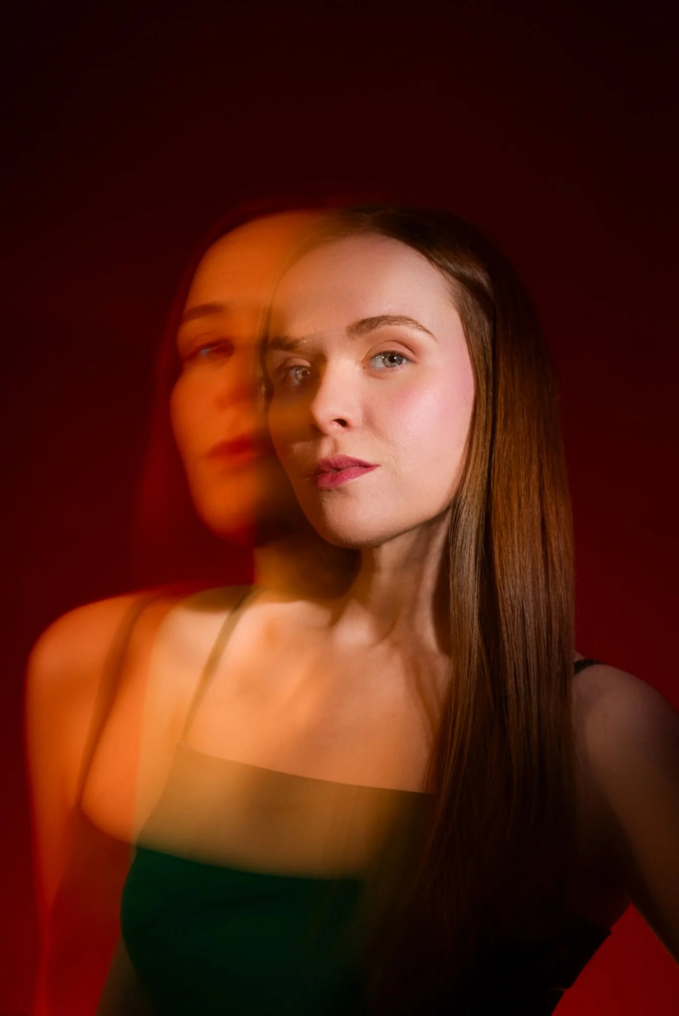 A portrait of a woman with long brown hair, wearing a black spaghetti strap top, against a dark red background, with a motion-blur effect creating a layered look.
