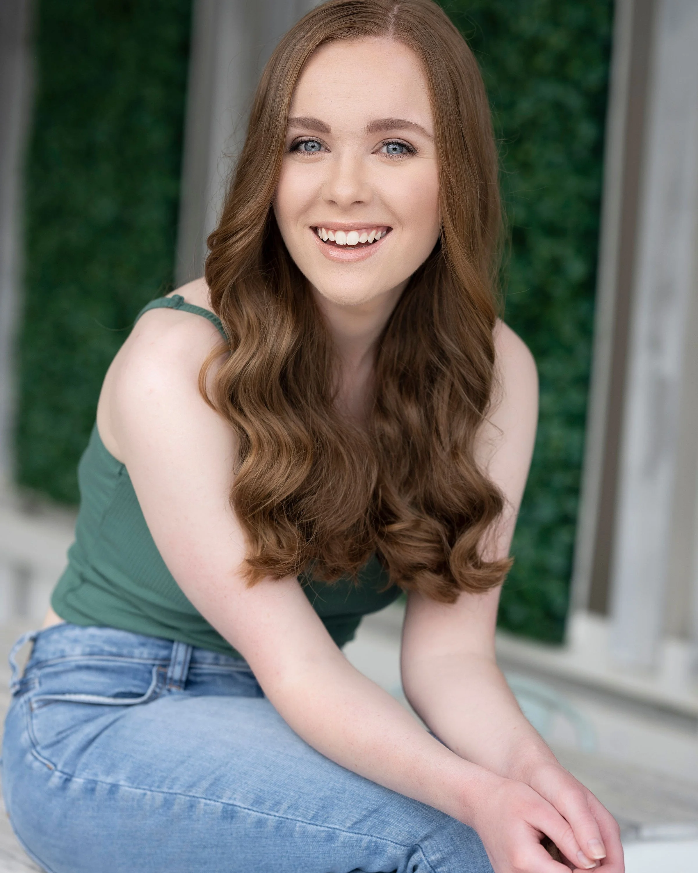 A young woman with long, wavy red hair, blue eyes, and fair skin, smiling while sitting outdoors in front of a green background.