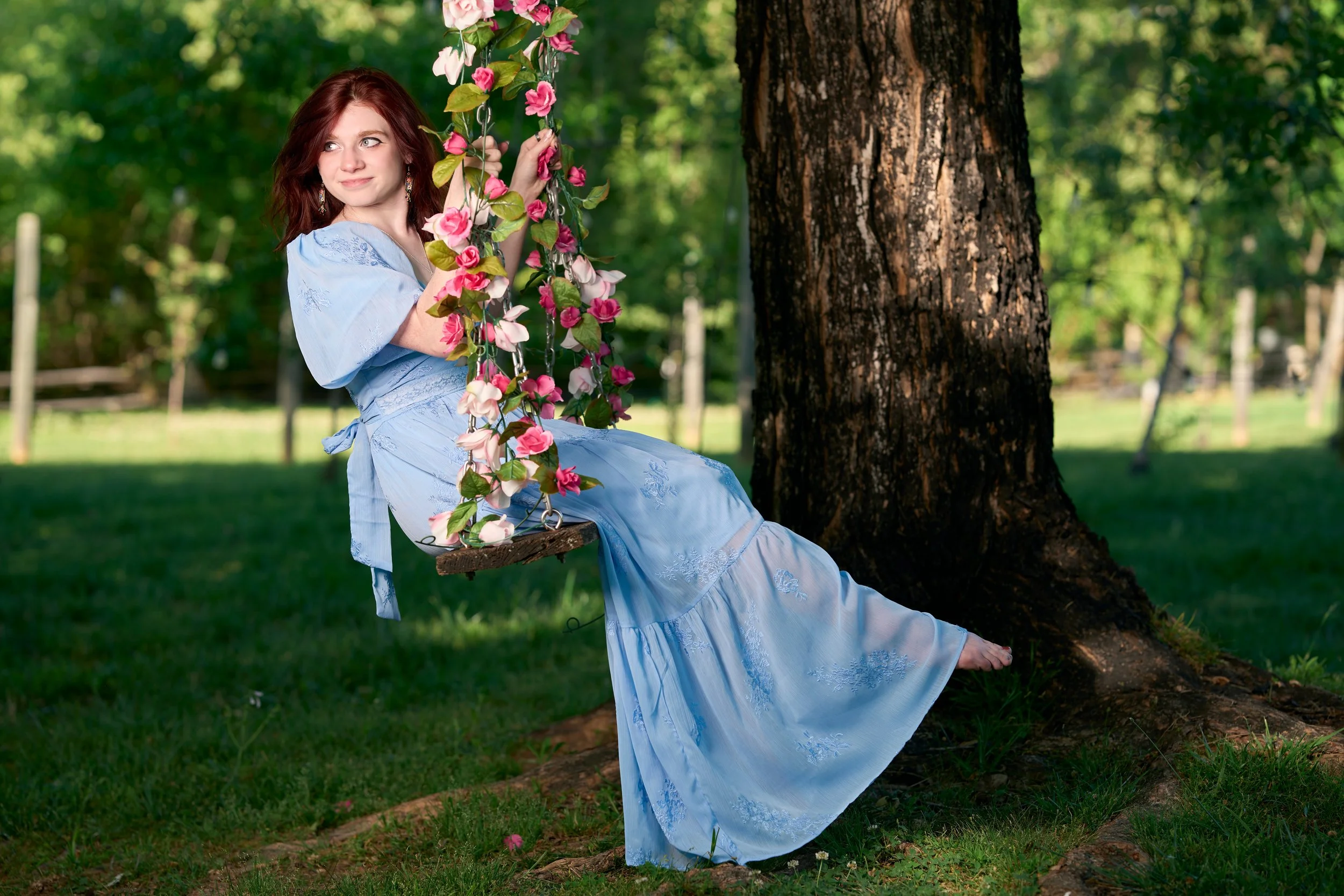 A woman in a light blue dress sitting on a wooden swing hanging from a tree in a lush green park. She has red hair and is holding onto the swing's chains, with pink flowers entwined with the chains.