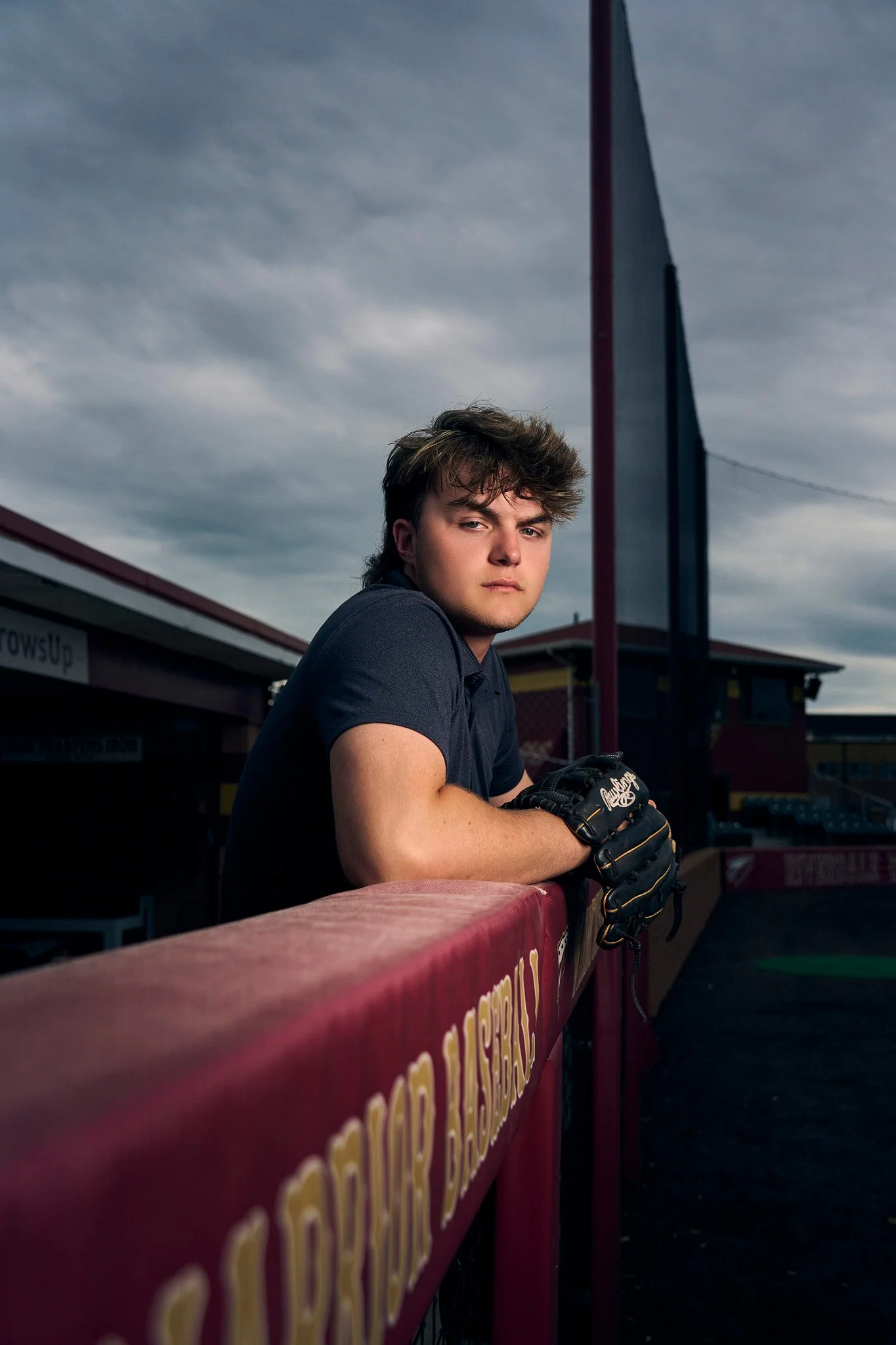 A young man with styled hair and a serious expression leaning on a red railing with yellow text, dressed in a dark T-shirt, wearing a baseball glove, at a baseball stadium under a cloudy sky.