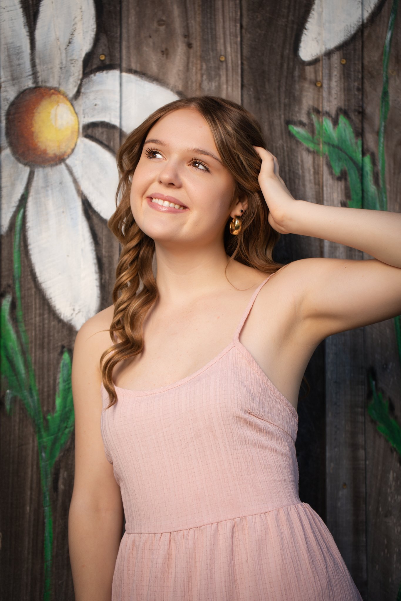 A young woman with long wavy brown hair, gold hoop earrings, and a pink sleeveless dress, smiling and looking slightly upwards, posing in front of a painted wooden wall with daisies.