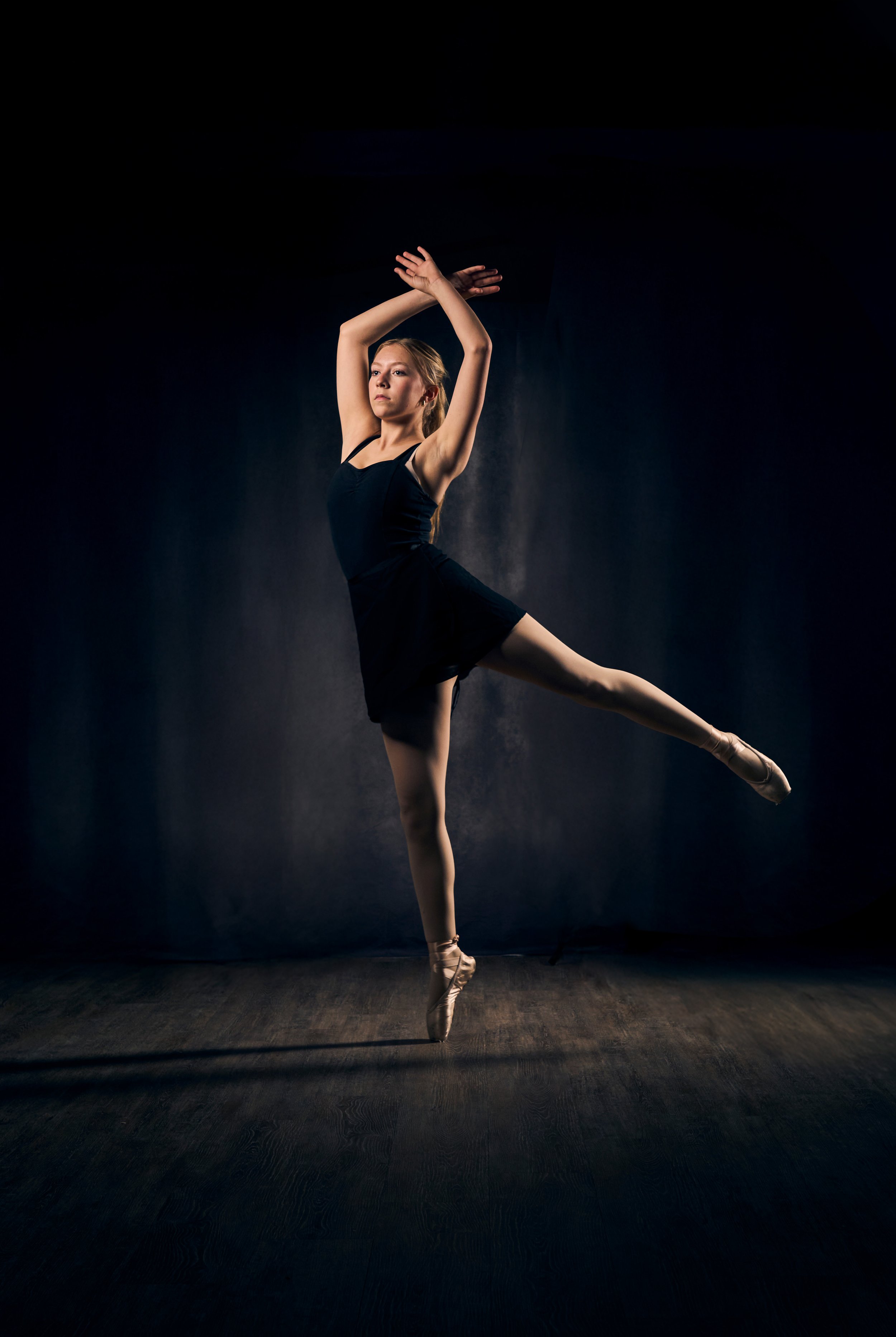 Ballet dancer performing on stage in a black leotard and pointe shoes, balancing on one leg with arms raised above head against a dark background.