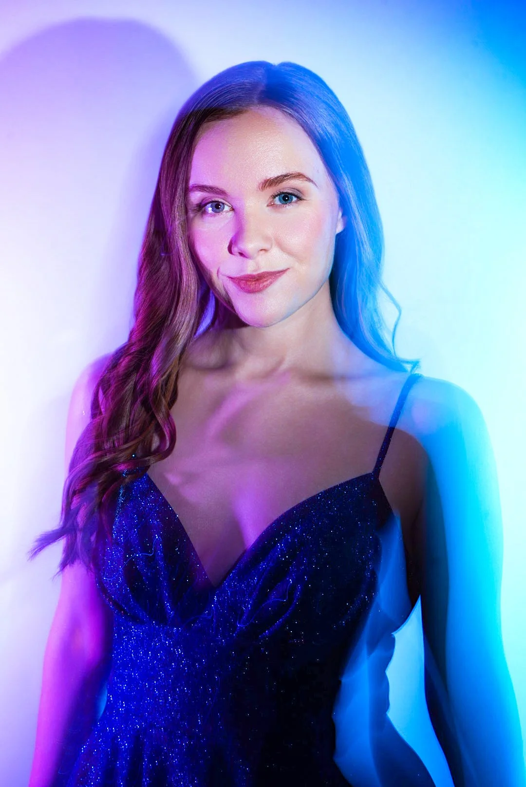 A woman with long wavy hair, wearing a dark sparkly dress with thin straps, standing against a white background illuminated with purple and blue lighting.