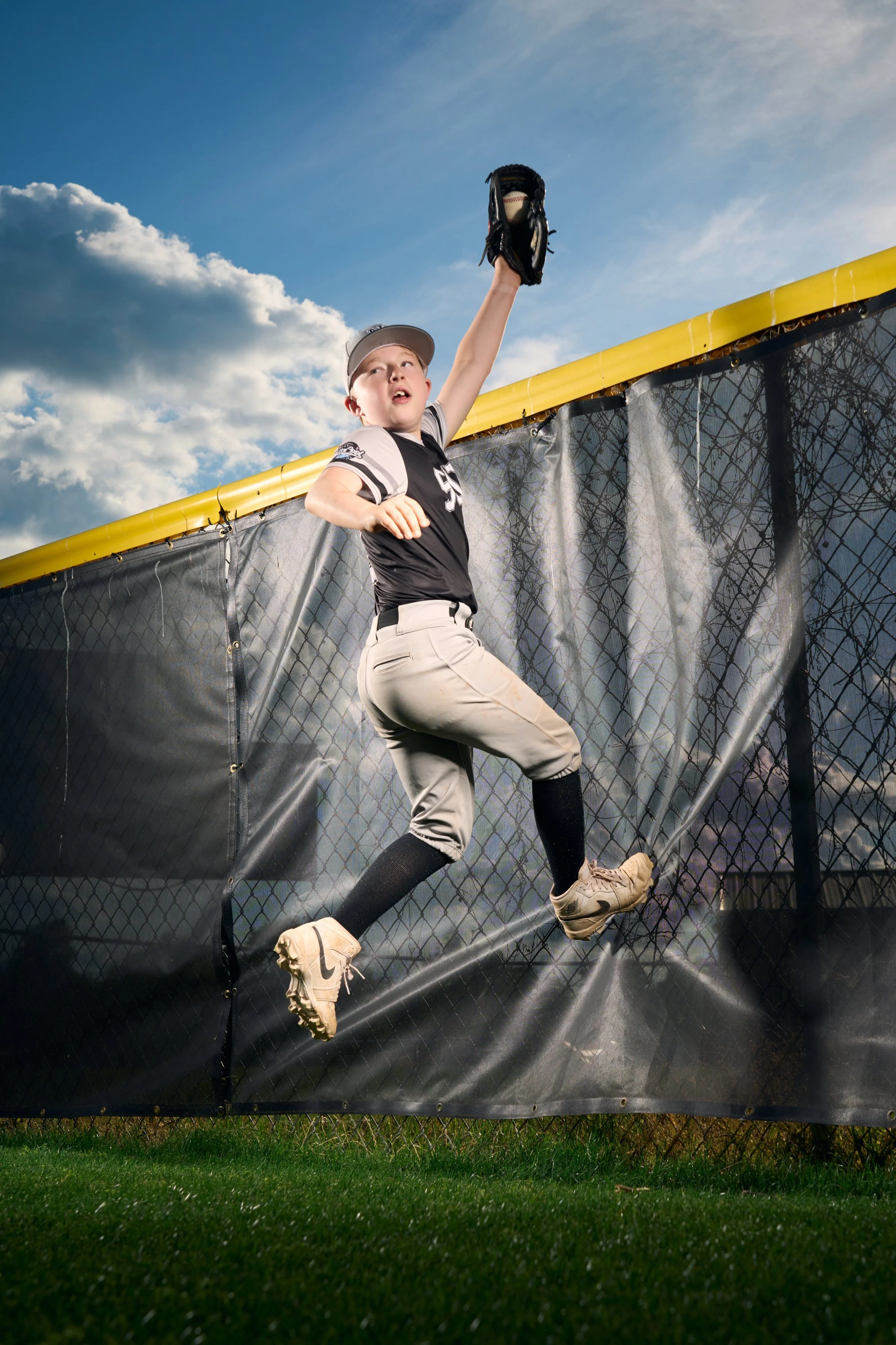 Young baseball player in mid-air catching a baseball with their glove during a game, with a chain-link fence and sky in the background.