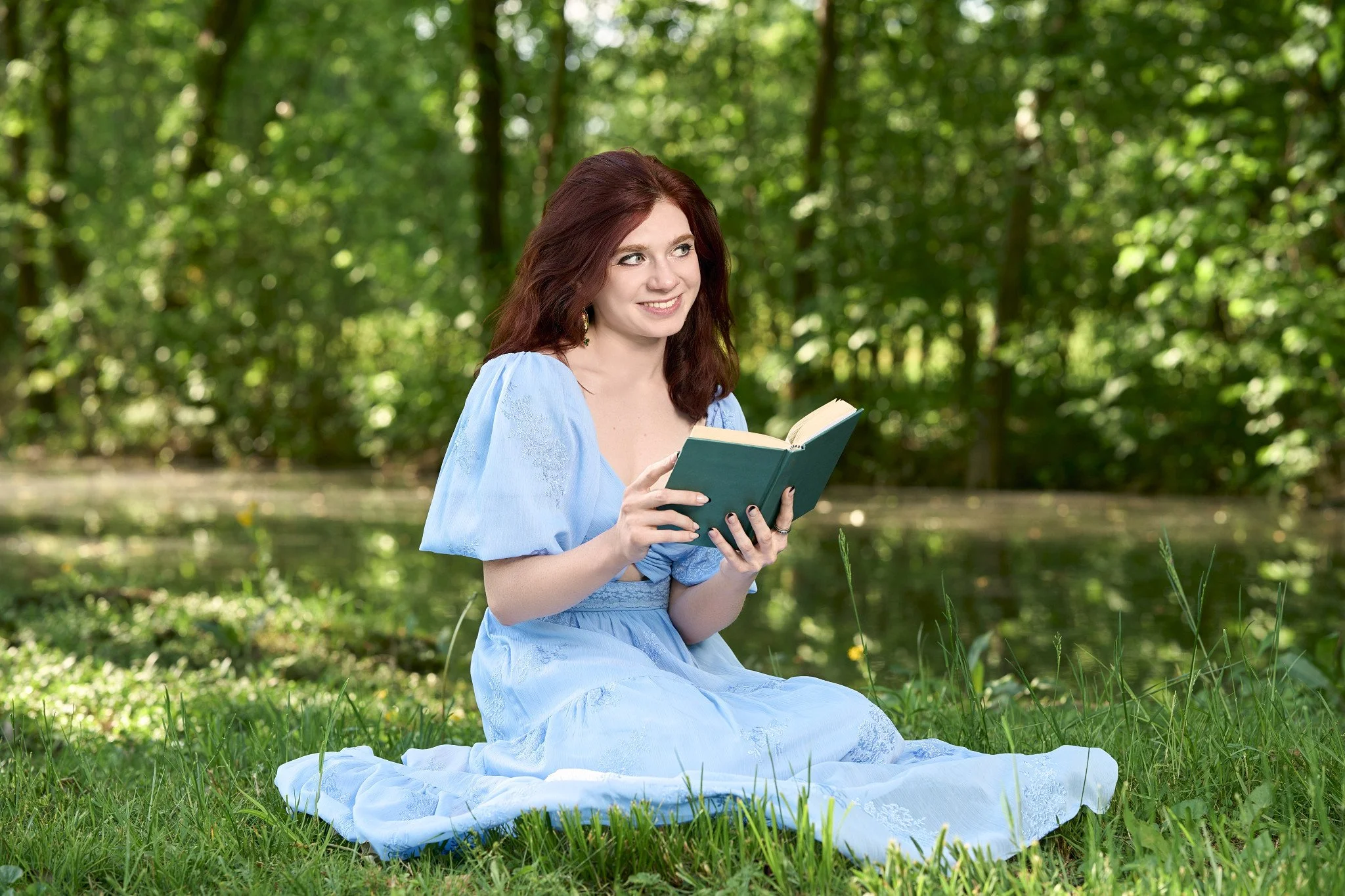 A woman with red hair wearing a light blue dress sitting outdoors on a grassy area, reading a book with trees in the background.