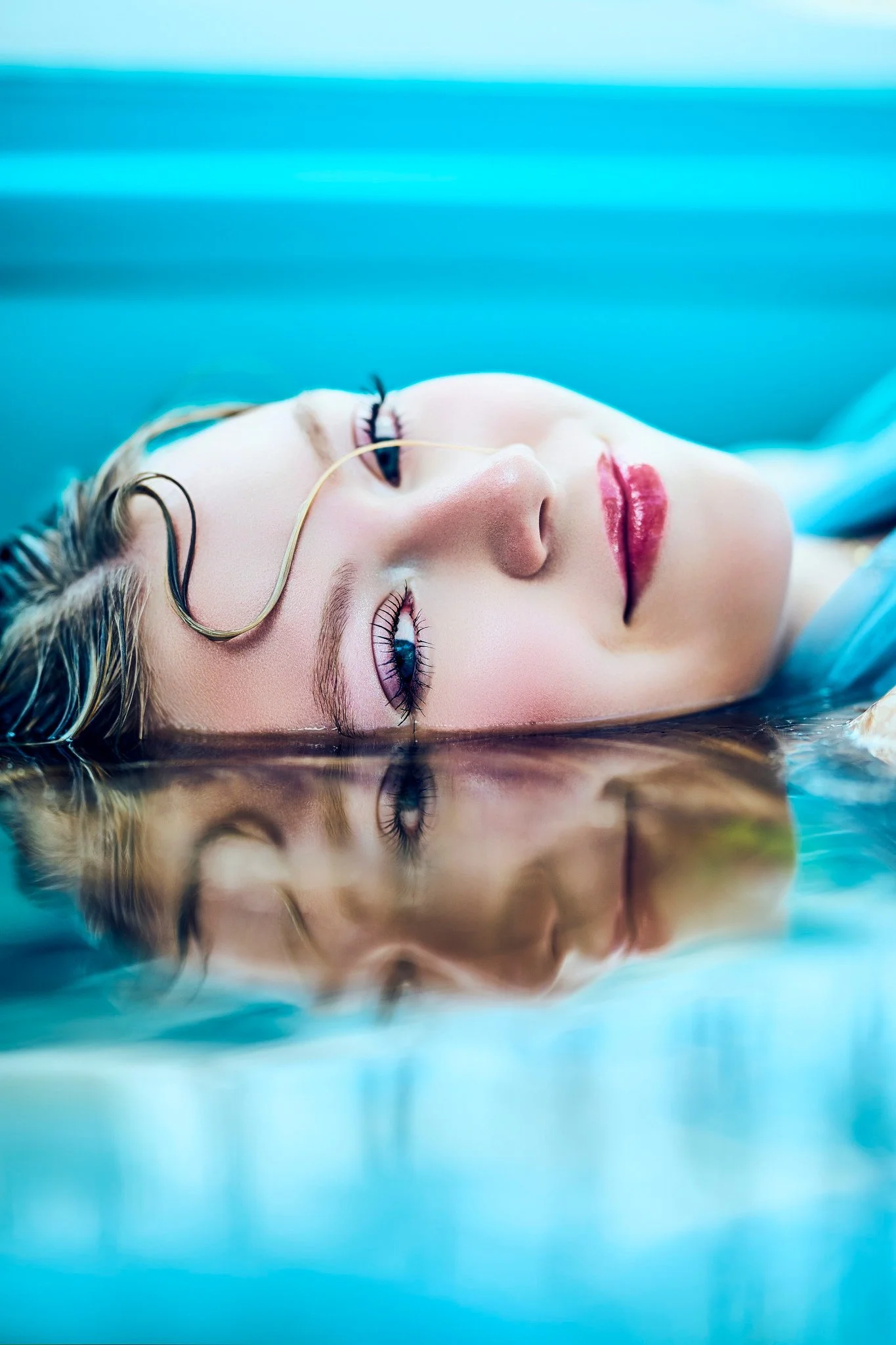 Close-up of a woman with blonde hair lying on her back in a blue environment, with a calm expression, closed eyes, and a reflective surface below her.