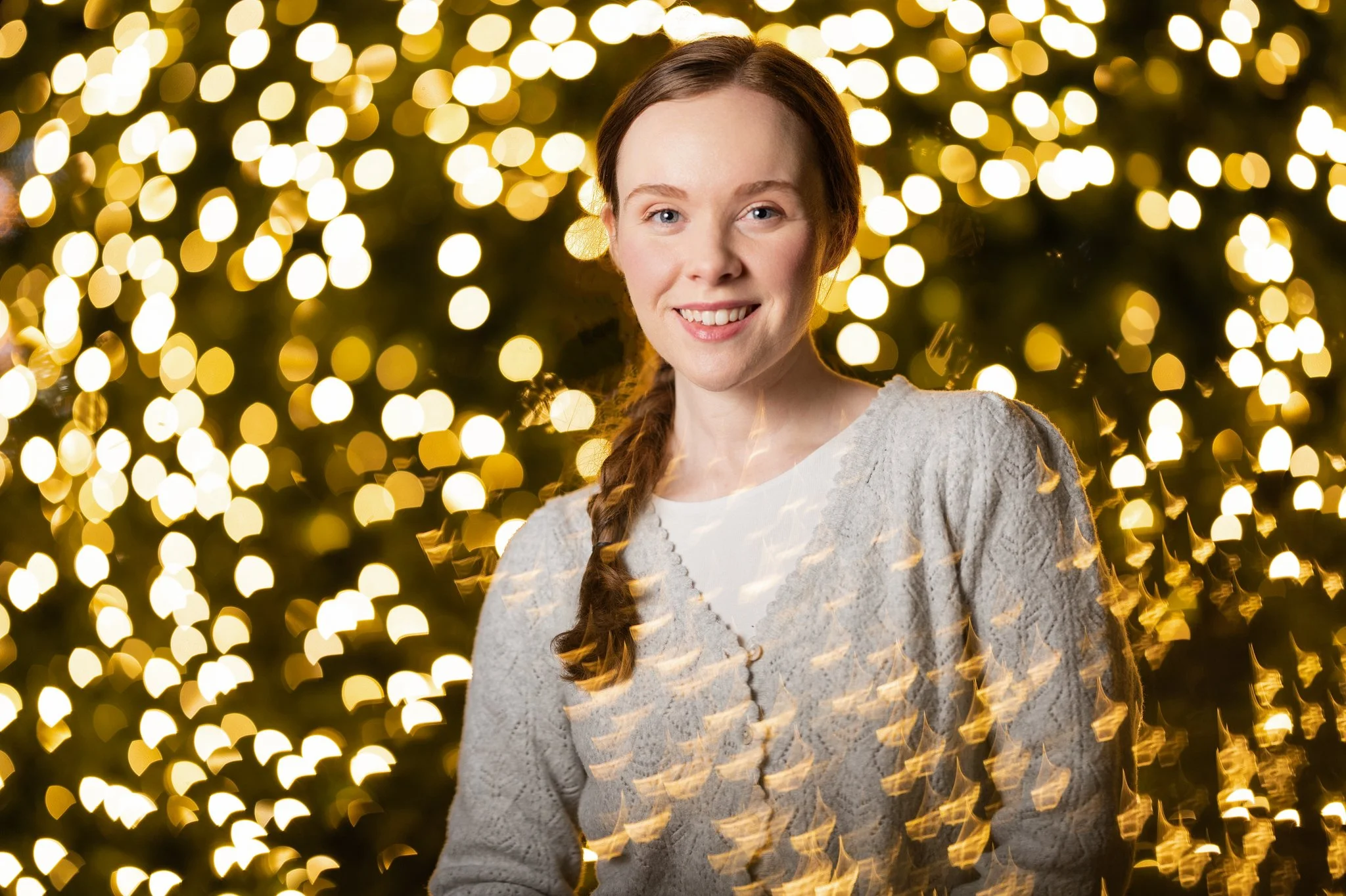 Young woman with long wavy hair and a white sweater smiling in front of a backdrop of blurred golden lights.