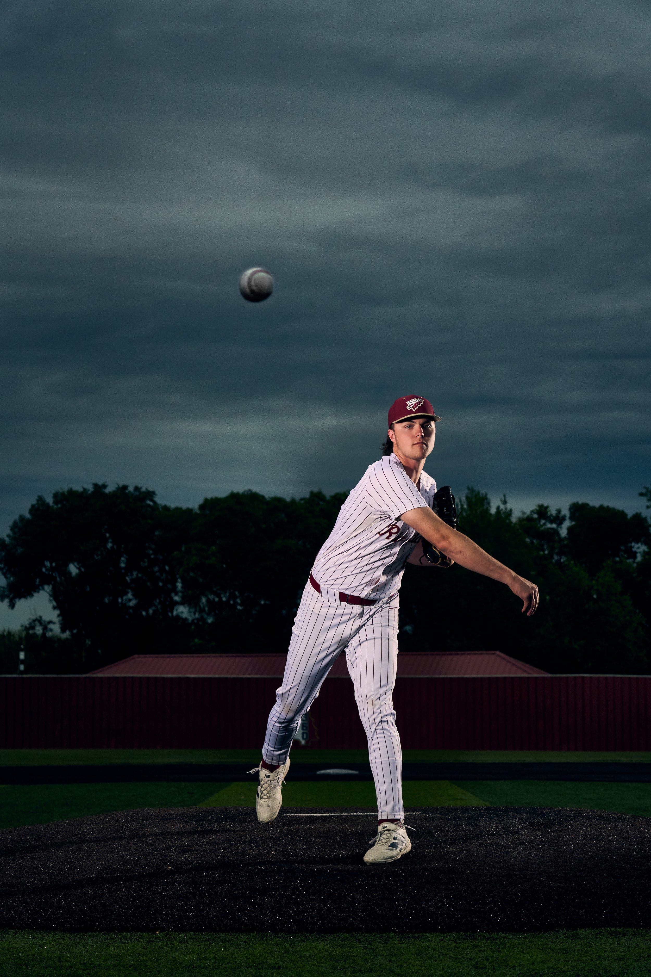 A baseball player in a uniform with pinstripes, wearing a maroon cap, is pitching on the mound at dusk or evening. The pitch is mid-flight toward the batter, with dark cloudy skies and trees in the background.