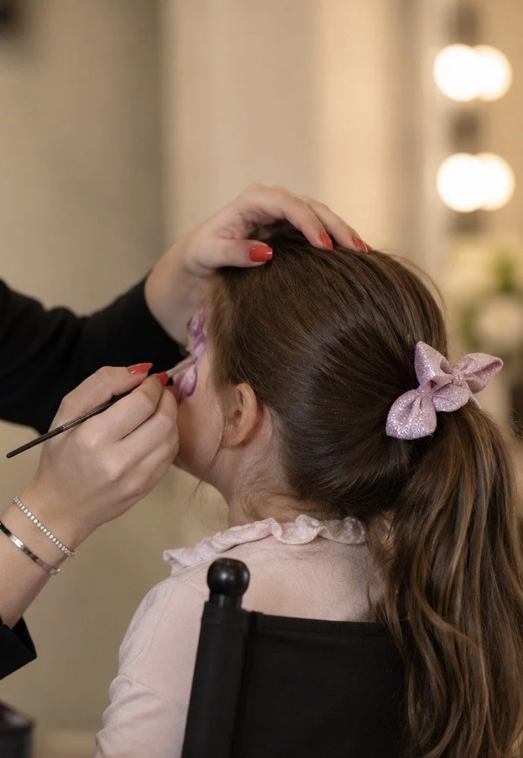 Une femme assise en fauteuil reçoit une séance de maquillage, avec de la peinture violette sur le visage, et porte un ruban rose dans ses cheveux bruns liés en queue de cheval.