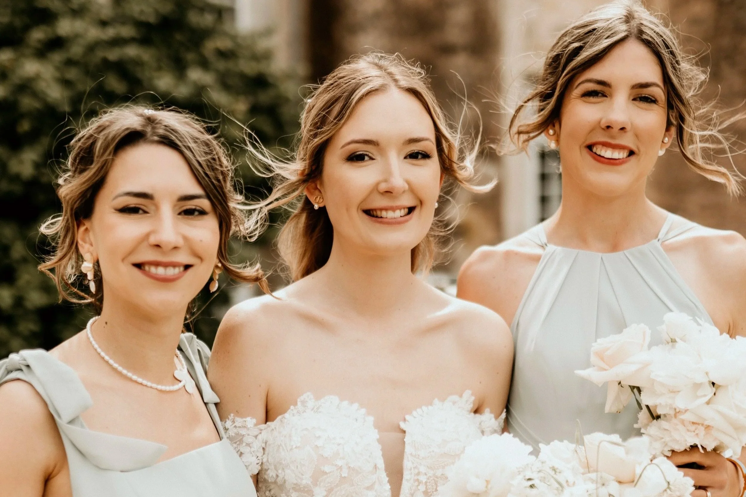 Trois femmes souriantes lors d'une cérémonie, habillées en robes de mariage et tenant des bouquets blancs, en plein air avec des arbres en arrière-plan.