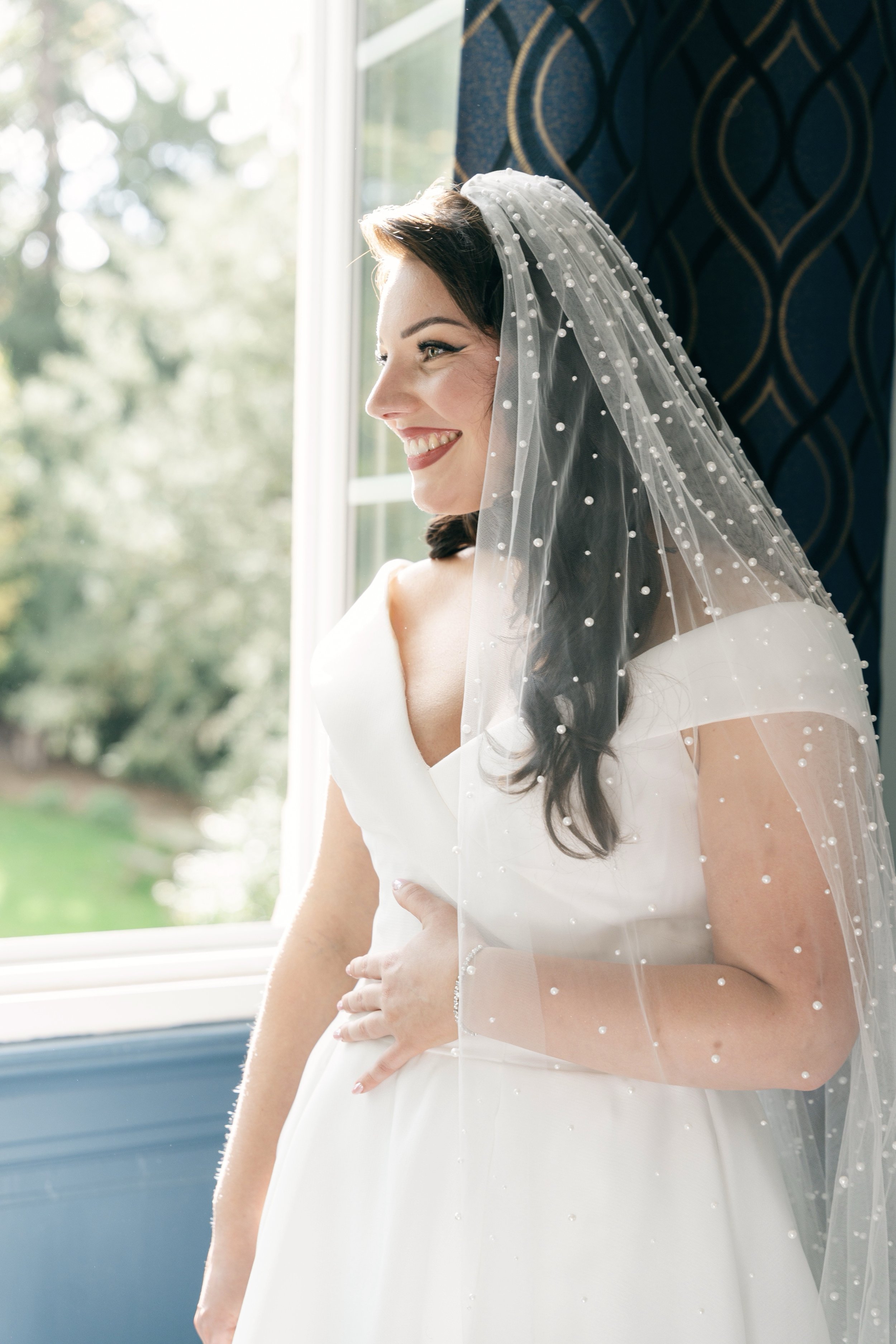 Une femme souriante en robe de mariée regarde par une fenêtre, avec un voile ornée de petits perles.