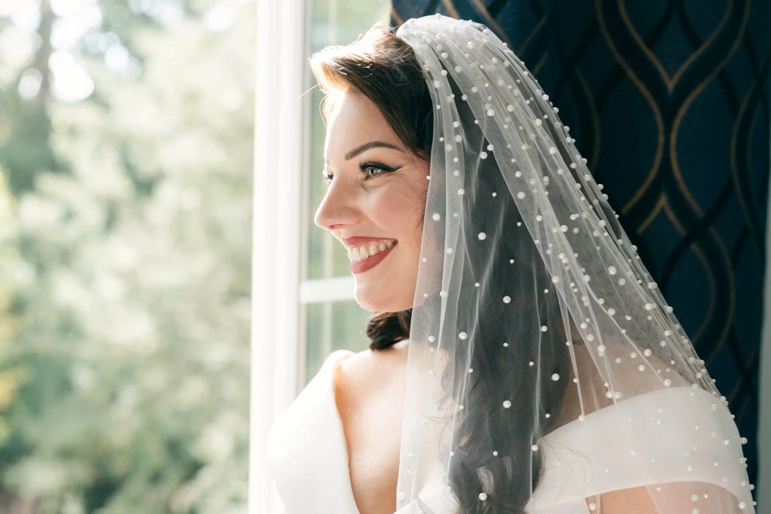 Une femme souriante porte un voile de mariée avec des perles, assise près d'une fenêtre laissant entrer la lumière naturelle.