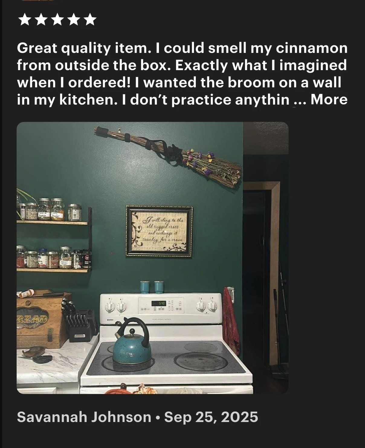 A kitchen with a green wall featuring a framed quote and a broom hanging above the stove. The stove has a teal kettle on it, and there are jars of spices on a wooden shelf. There is also a knife block and a cutting board labeled 'bread' on the counter.