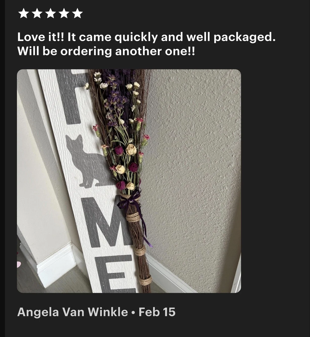 A decorative sign with a cat silhouette in black and large letters reading 'HOME', leaning against a wall. Attached to the sign is a bundle of dried flowers with purple, beige, and pink blossoms, tied with purple string.