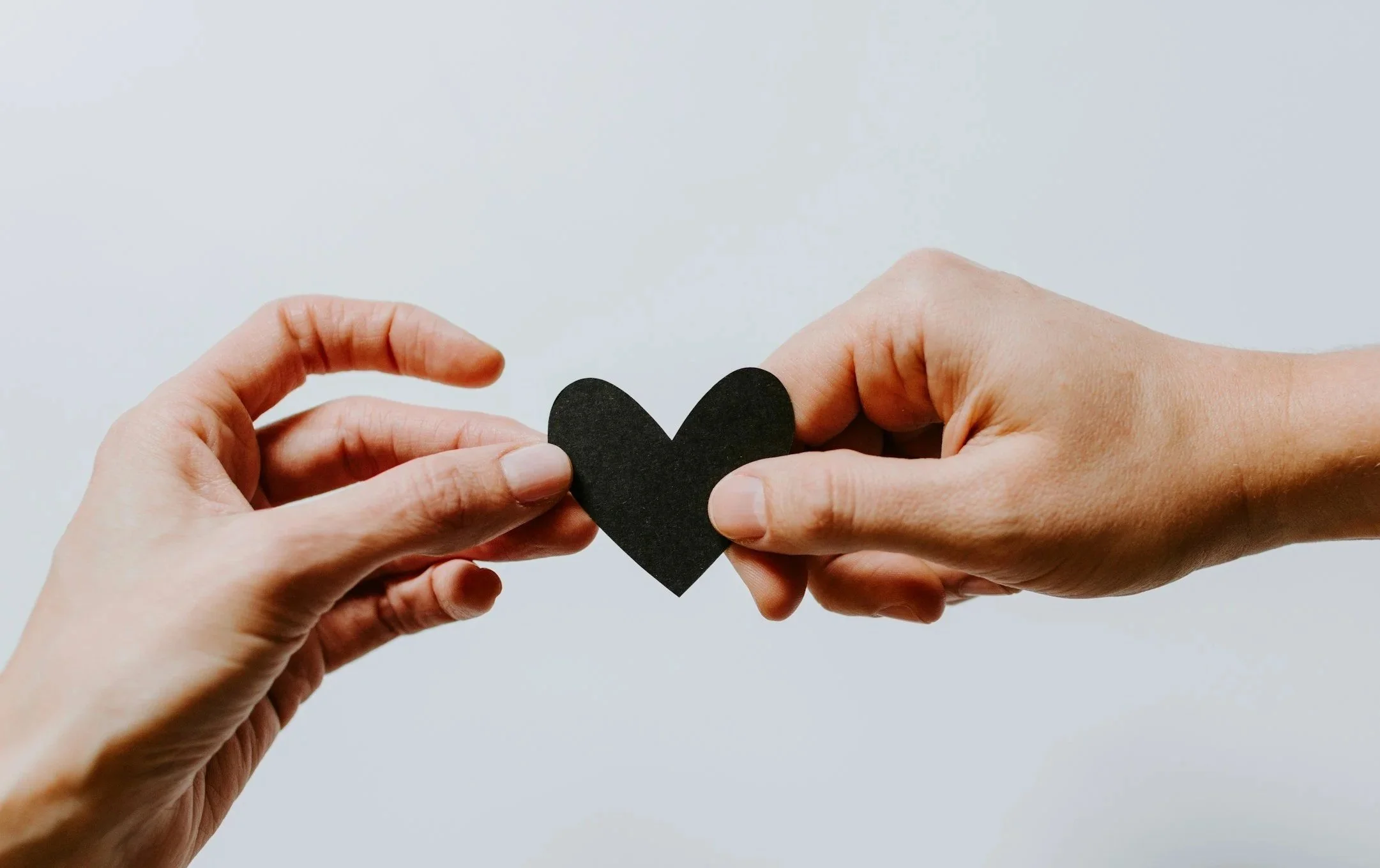 Two pairs of hands holding a black paper heart against a light grey background.