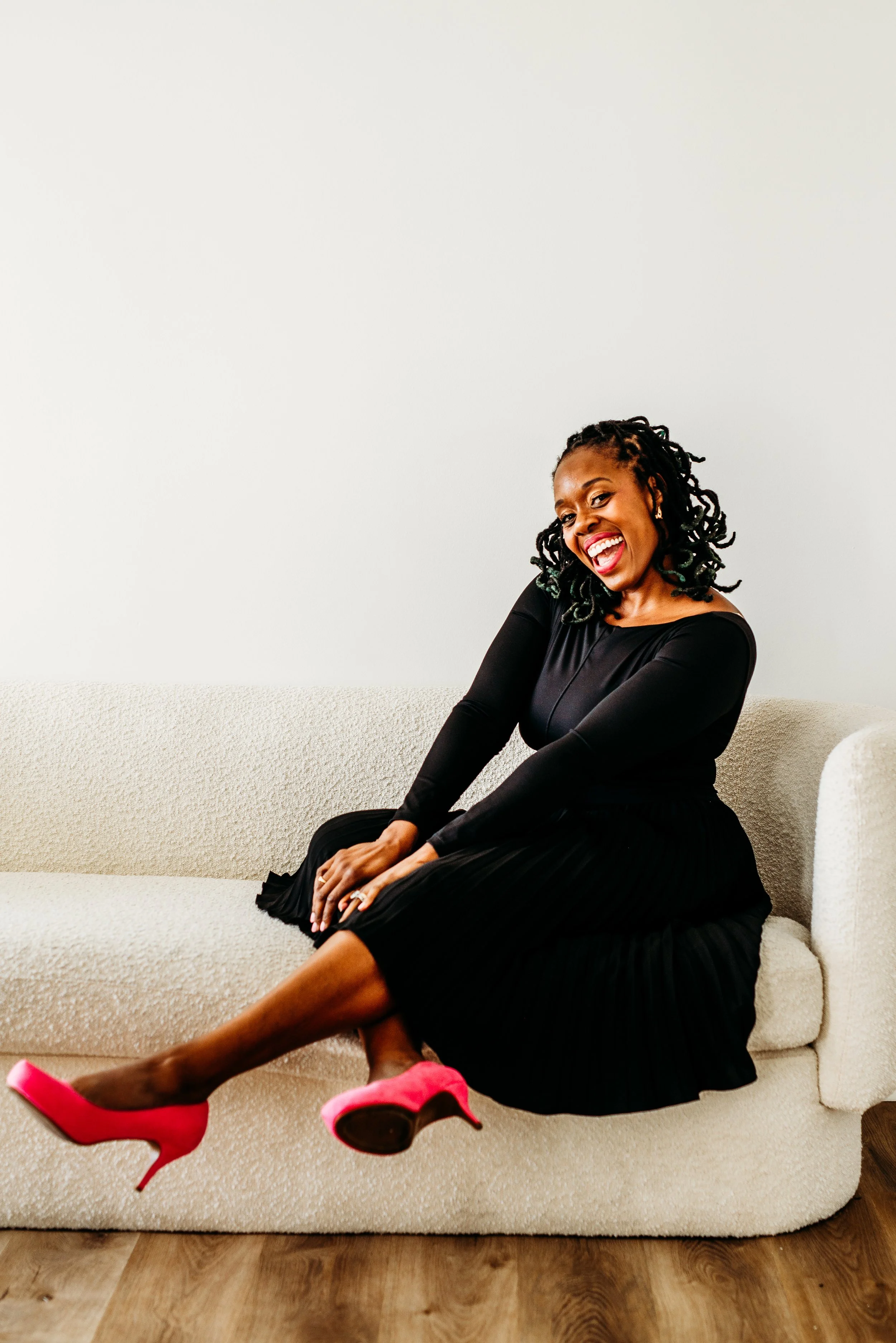A smiling woman with curly hair wearing a black dress and red high heels, sitting on a cream-colored sofa against a white wall.