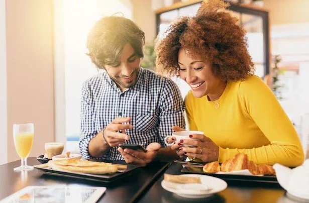 Two friends enjoying a meal at a restaurant, looking at a smartphone and laughing.