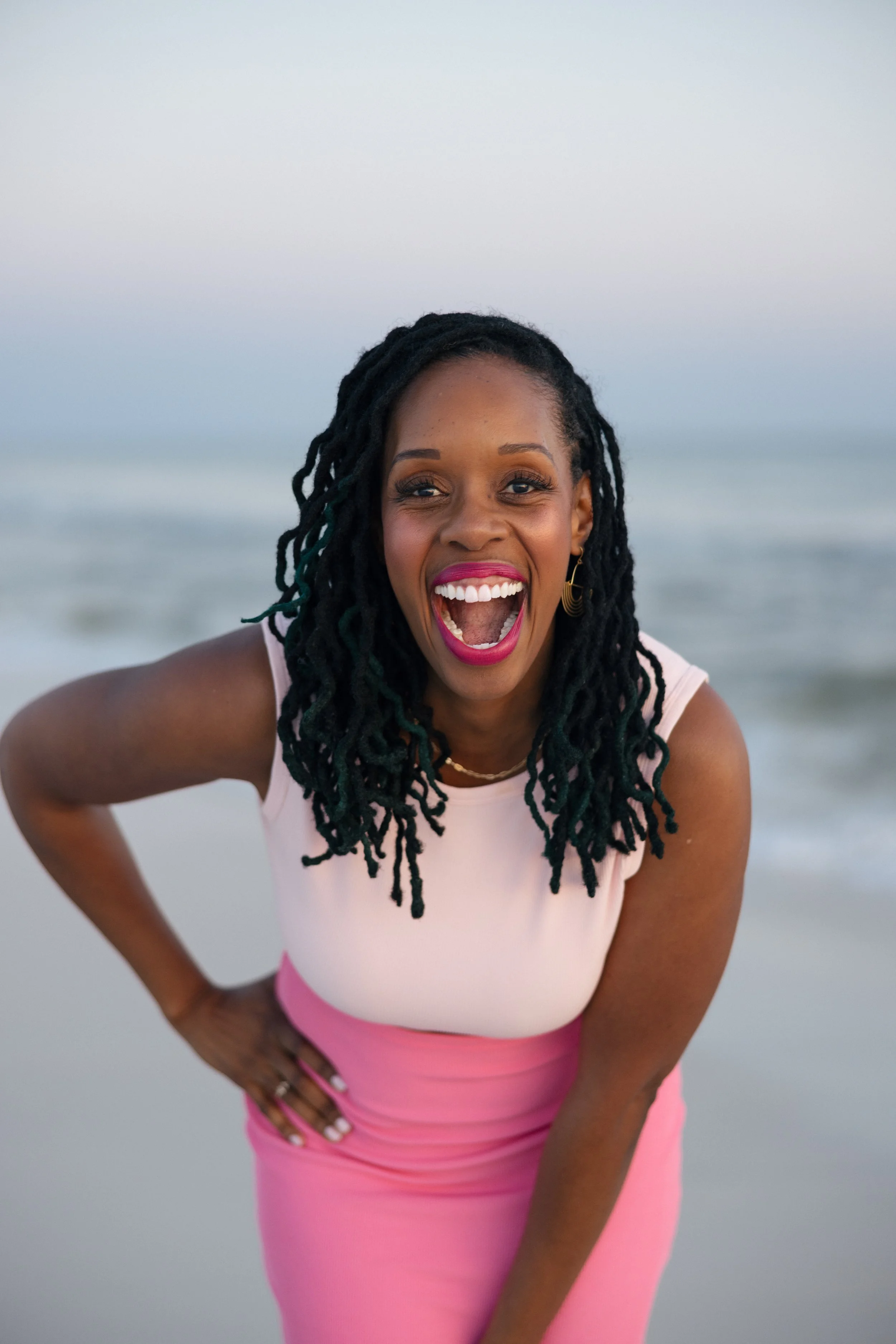 A woman with long black dreadlocks and dark skin curiously leaning forward on a beach, wearing a white and pink outfit, smiling widely with red lipstick.