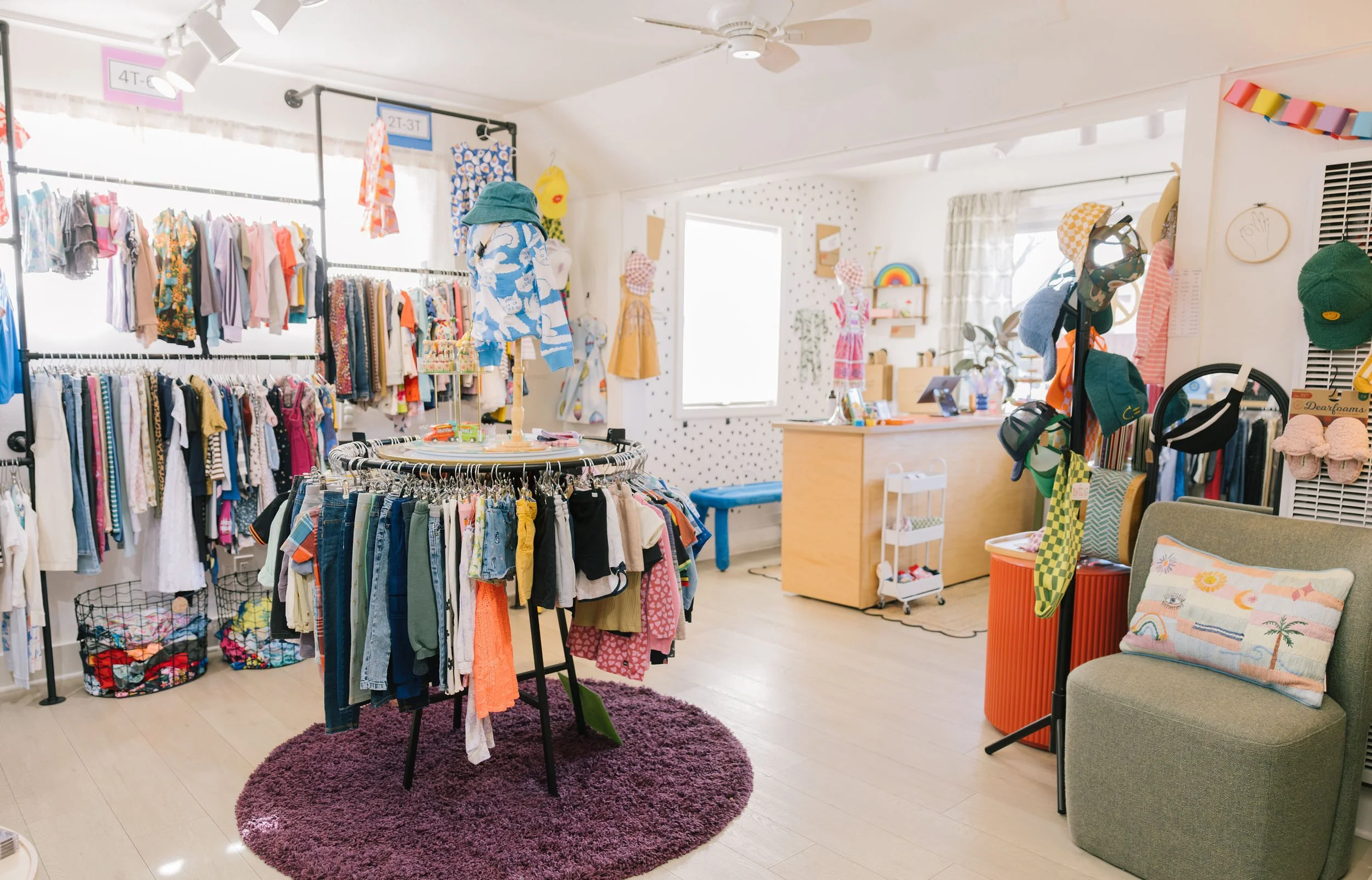 Interior of Hand It Down Kid to Kid Resale Shop in Santa Cruz, CA — a children's clothing store with racks of colorful clothing, hats, and accessories, a small desk, and a purple rug on the light-colored floor.