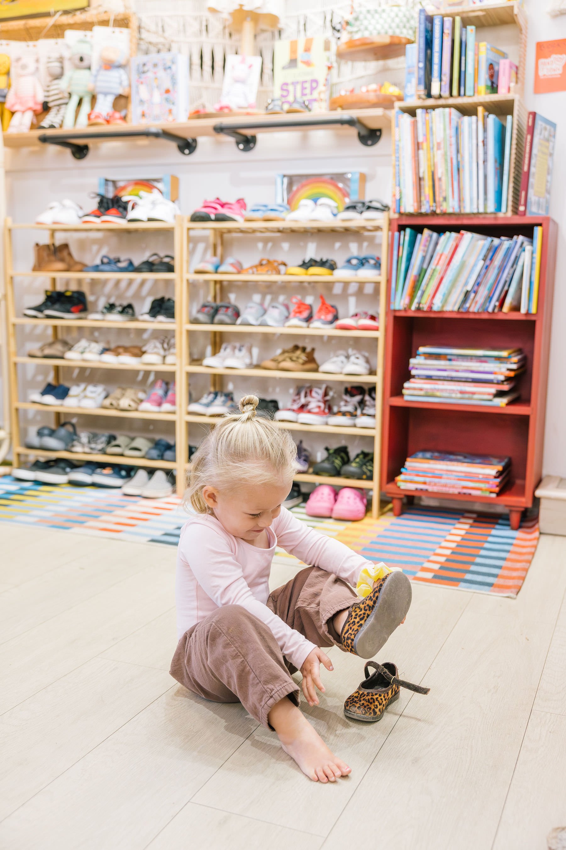 A young girl is sitting on the floor of a store or library, putting on her shoe. Behind her, there are shelves filled with colorful shoes and books, and a patterned rug on the floor.