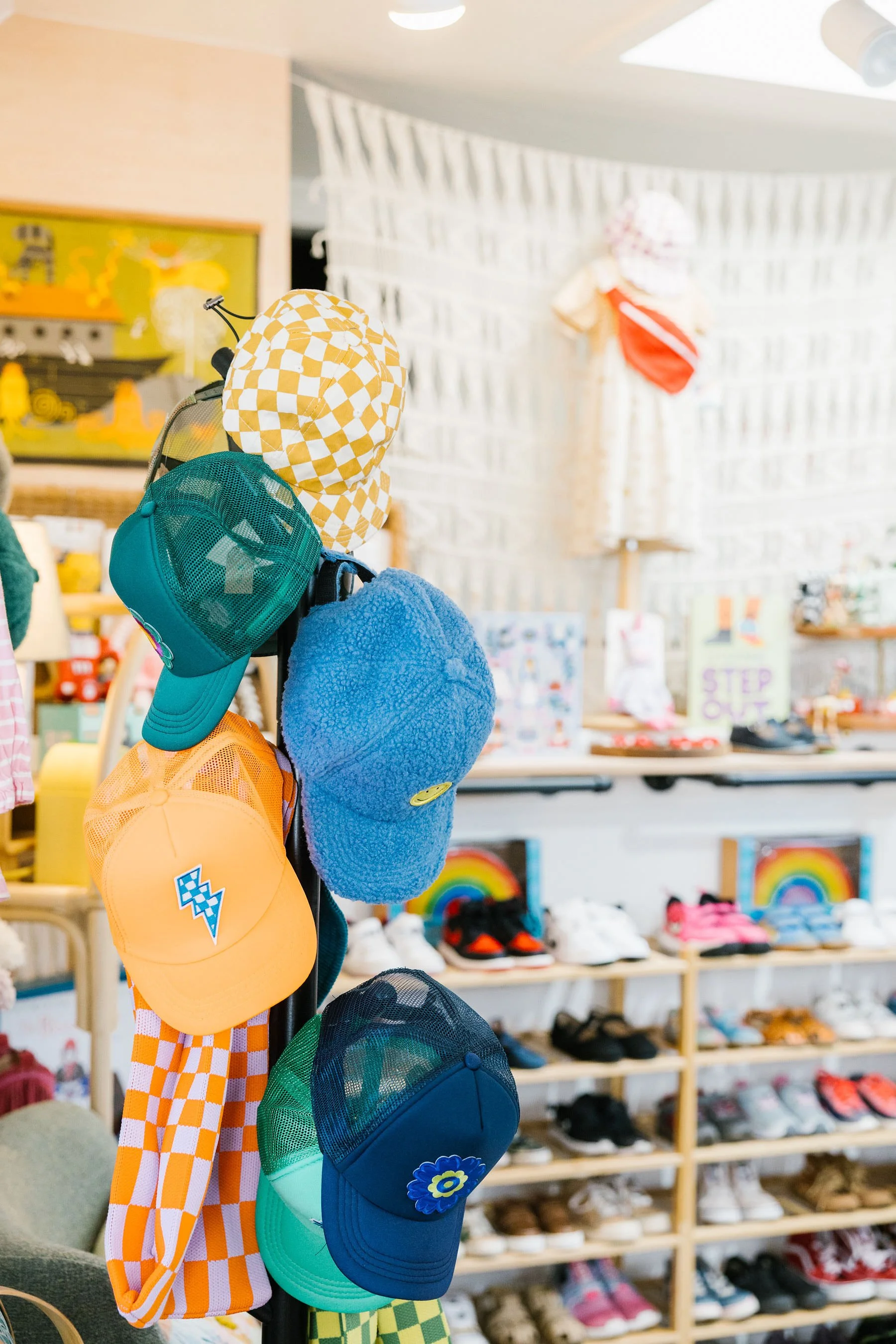 A display of colorful children's hats on a stand inside Hand It Down Kid's Resale store in Santa Cruz, CA, with a background of shelves filled with shoes and other second-hand kid's accessories, toys, books, and more.