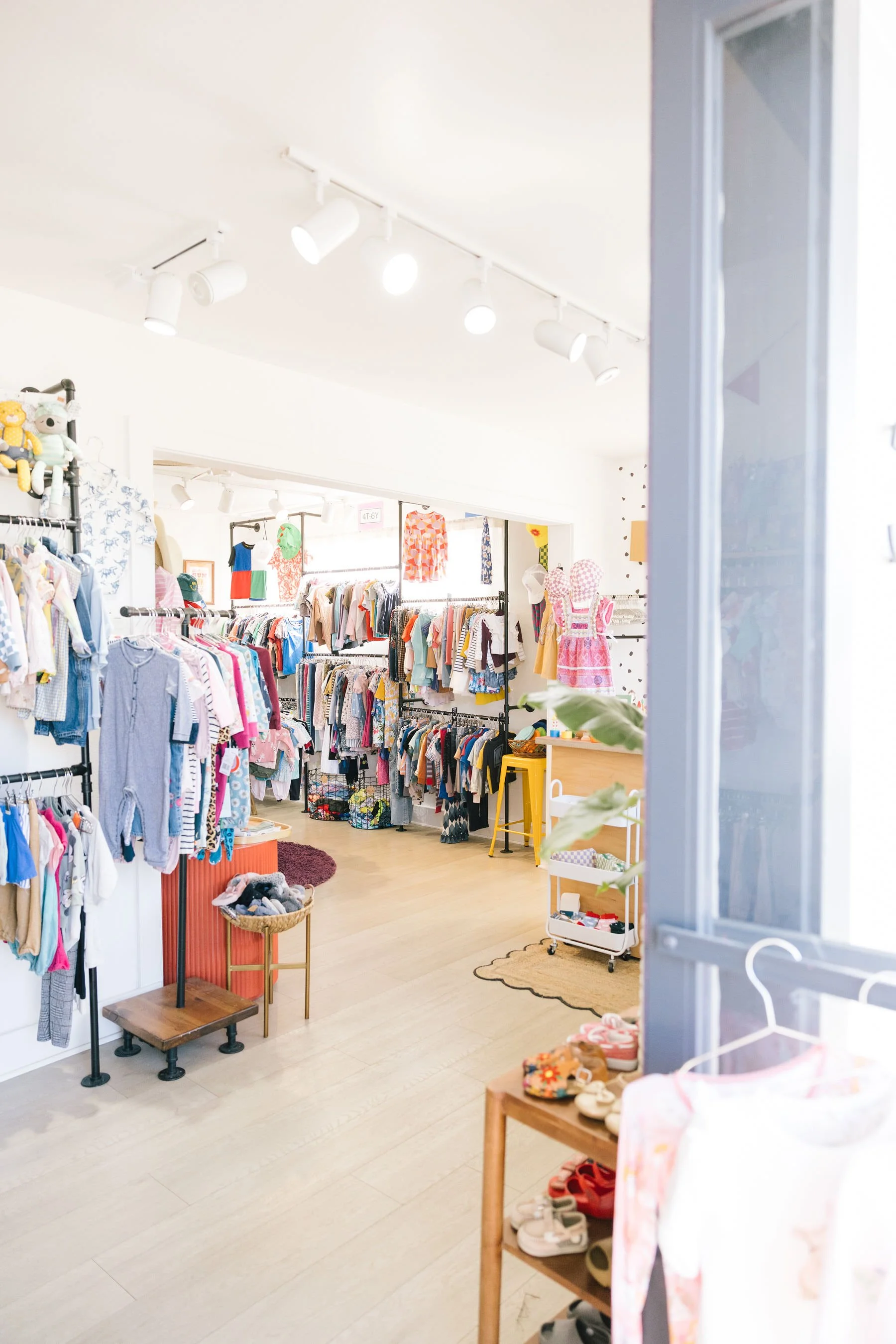 Clothing store with racks of children's clothes, shoes on a small shelf, and soft toys on a wall shelf, seen through a partially open door.