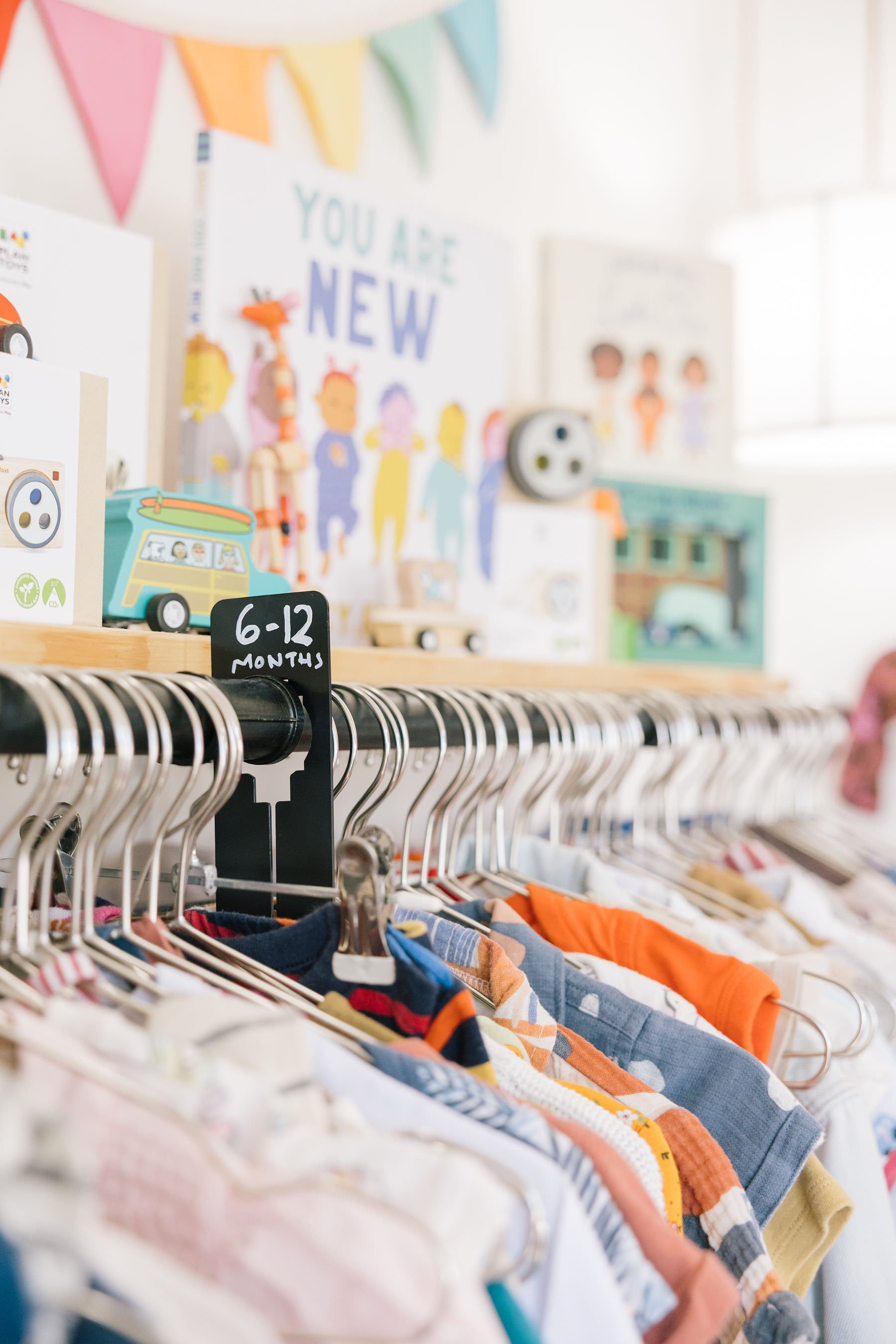Clothes for children, size 6-12 months, hanging on a rack in a store, with colorful shirts visible in the foreground.