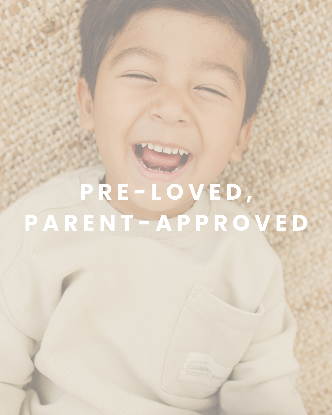 A young boy with short dark hair and a big smile showing missing front teeth, lying on a textured surface, wearing a light-colored sweatshirt. Text overlay says 'Pre-Loved, Parent-Approved.'