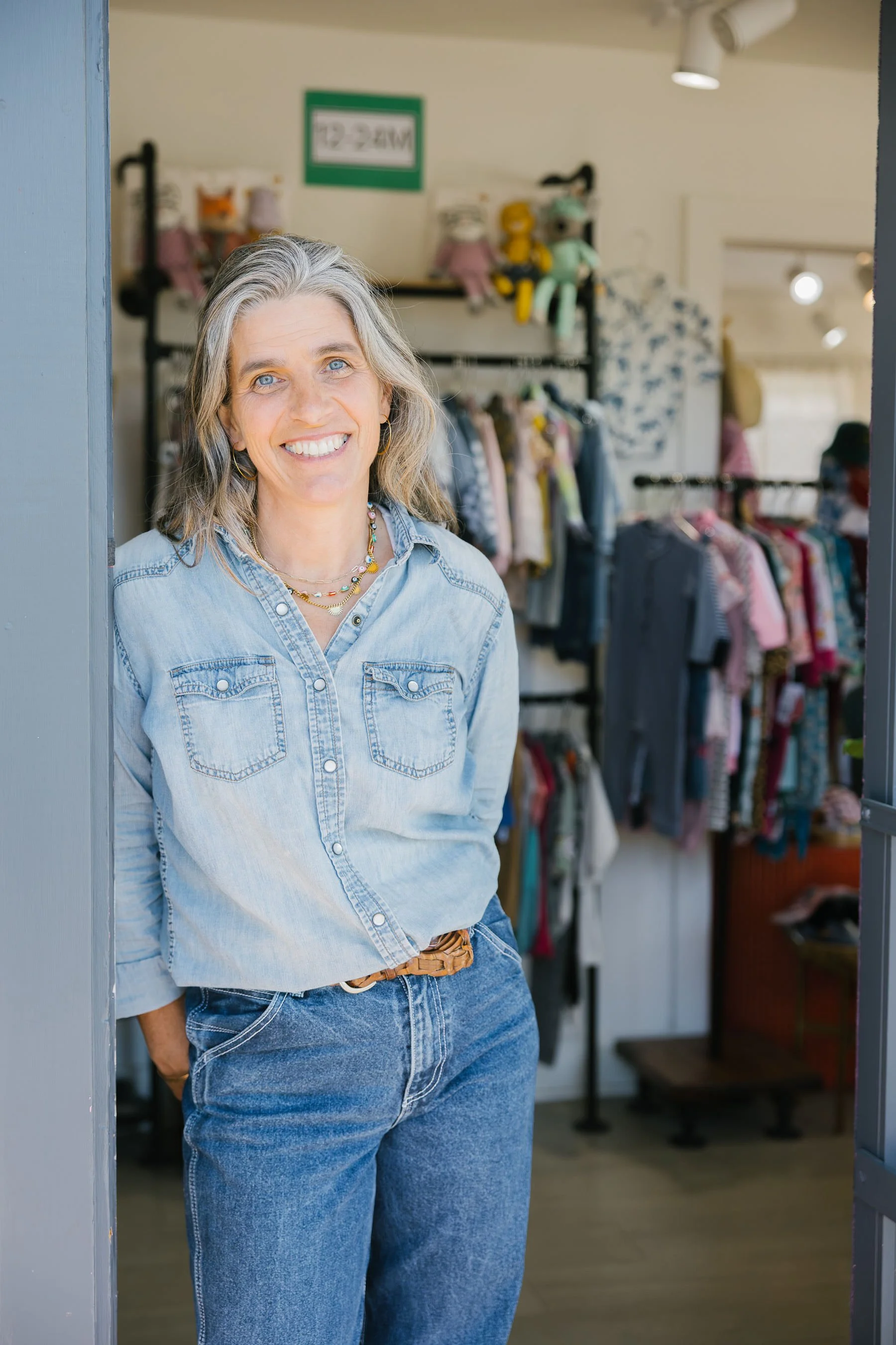 Smiling headshot of Hand It Down Kid's Resale Shop owner, Tori Koneya, wearing an all denim outfit, leaning in the doorway of her first Pleasure Point, Santa Cruz, CA storefront.