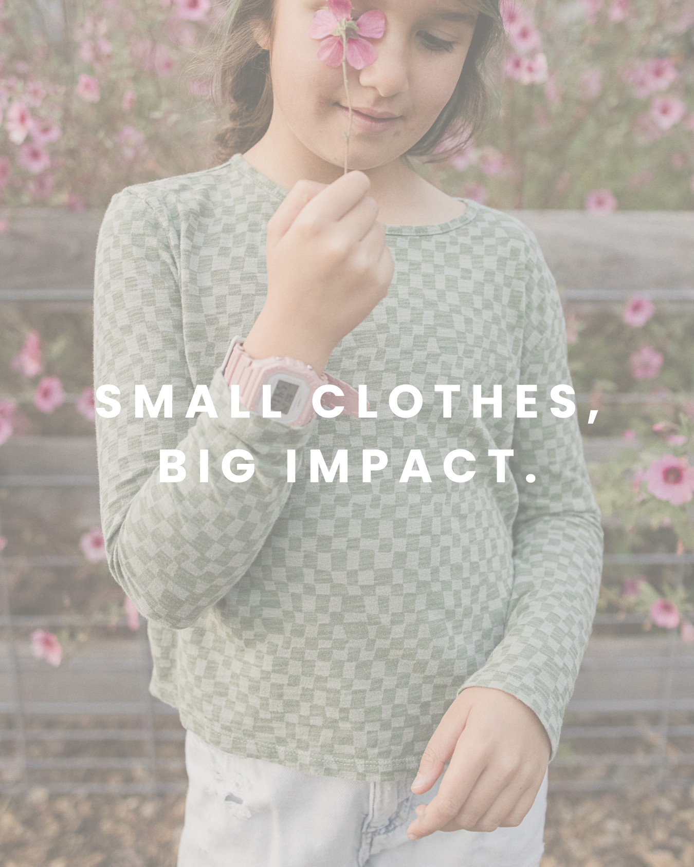 A little girl standing in front of a wood fence with pink flowers, holding a flower close to her face with a gentle smile and a watch on her wrist. She is wearing a light checkered blouse and text that says "Small Clothes, Big Impact" is laid on top.