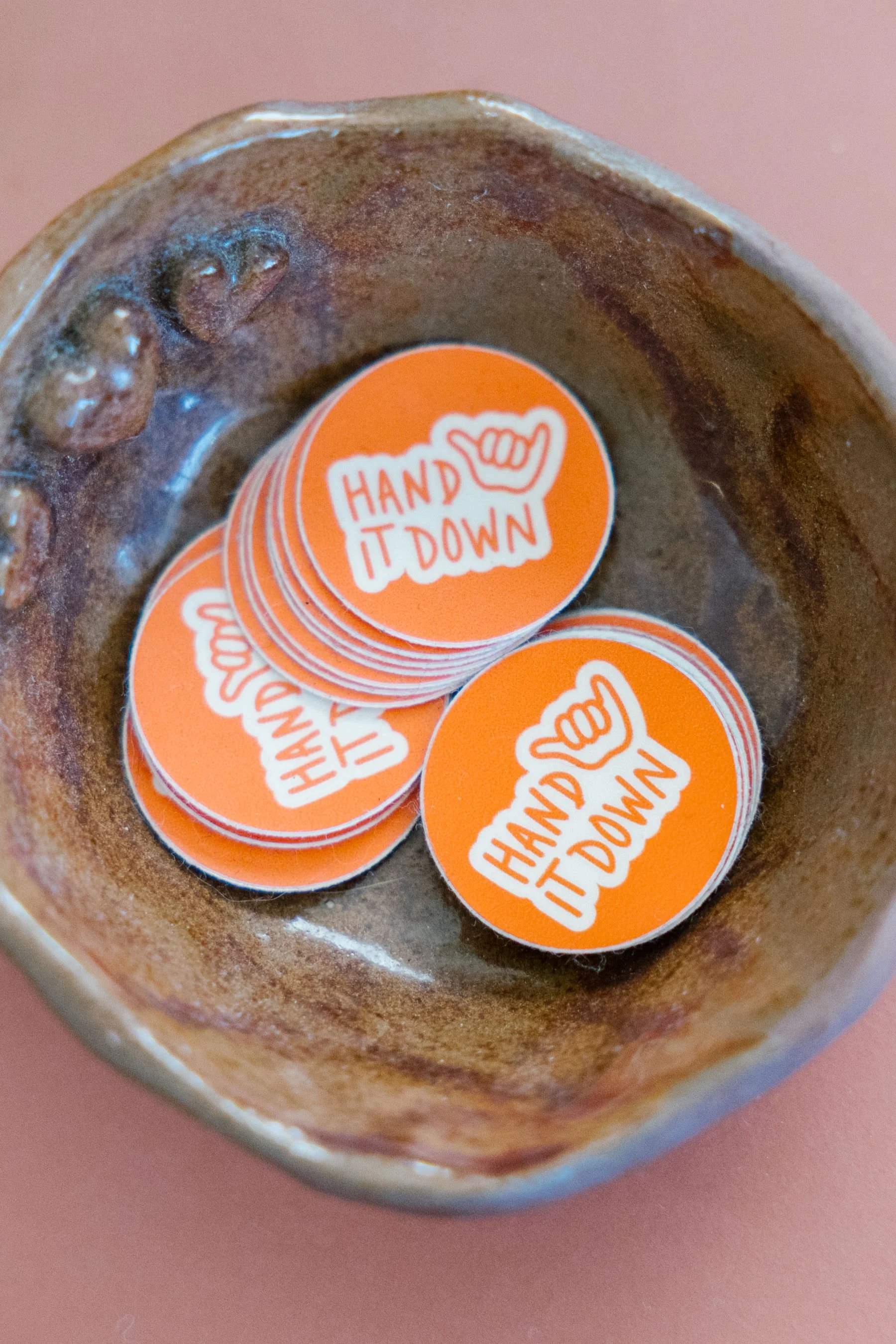 A close-up view of the bottom inside of a ceramic bowl, filled with numerous orange stickers that have a white outlined hand making a shaka sign and the text "HAND IT DOWN".