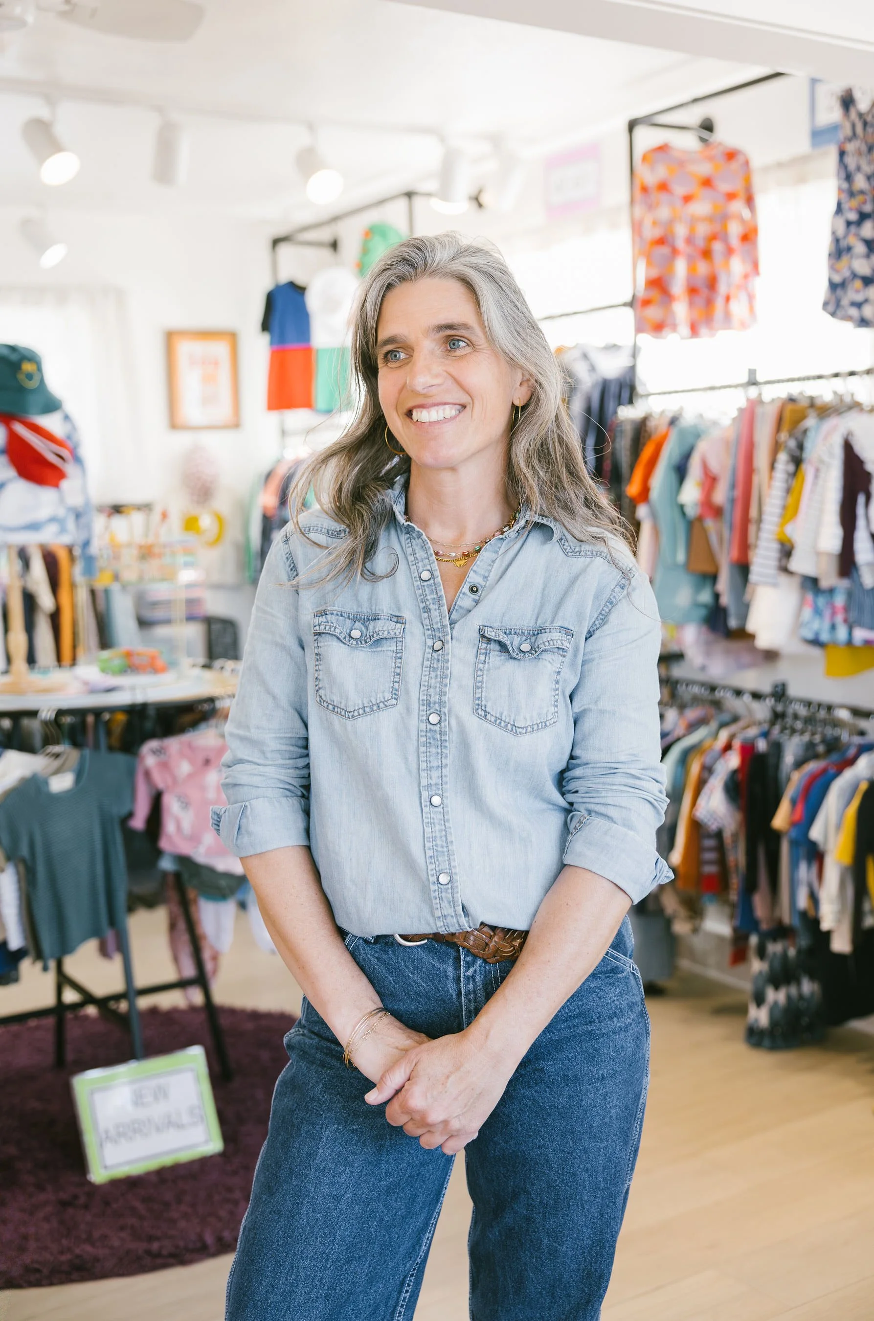 A woman with gray hair and blue eyes smiling, standing inside a colorful clothing store. She is wearing a light denim shirt and jeans, with clothing racks and shelves full of kids' clothing in the background.