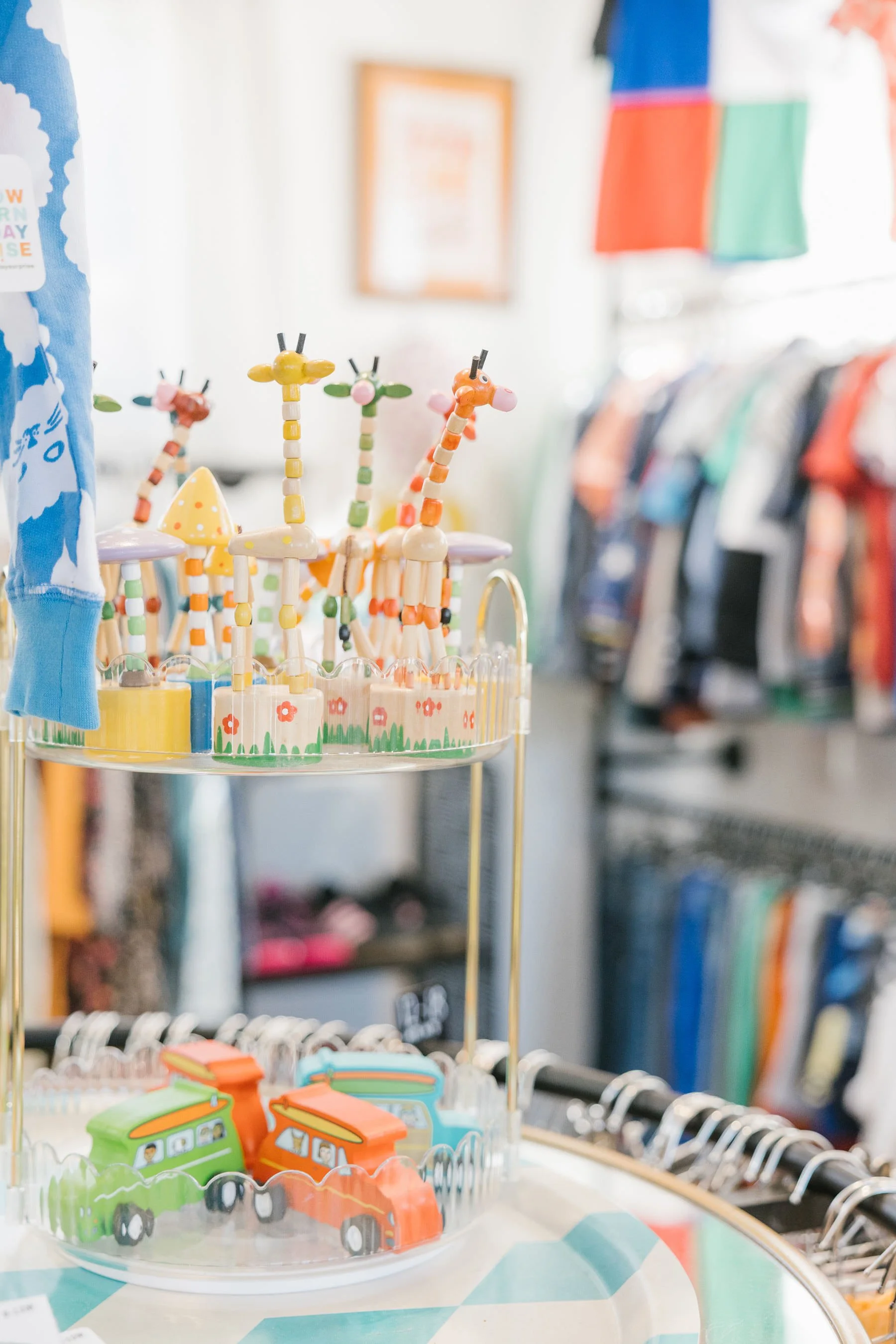 Colorful wooden giraffe toys arranged on a three-tier display stand in a store, with clothing racks and other blurred merchandise in the background.