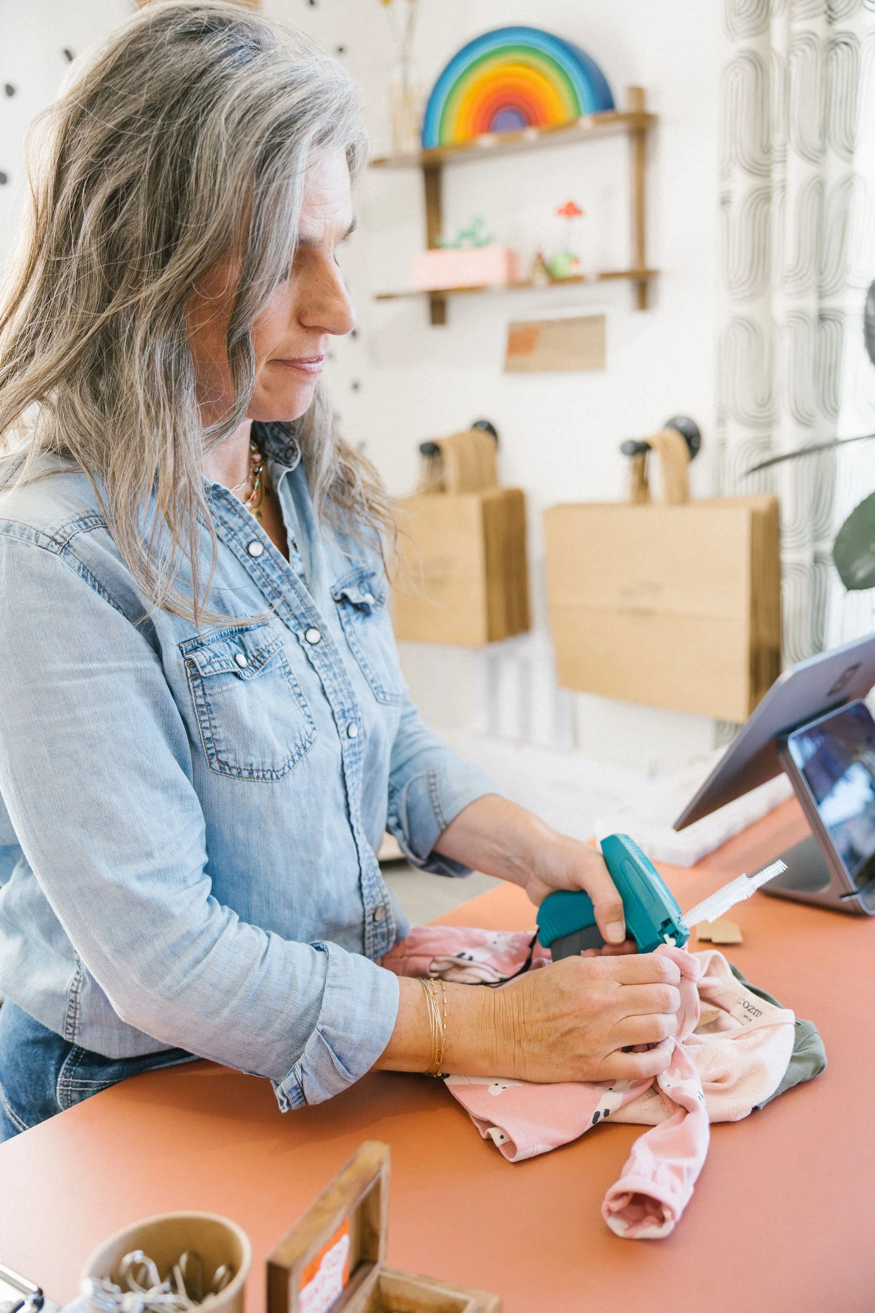 A woman in a denim shirt using a handheld pricing gun to tag a piece of children's clothing at Hand It Down Kid's Resale Shop in Santa Cruz, CA.