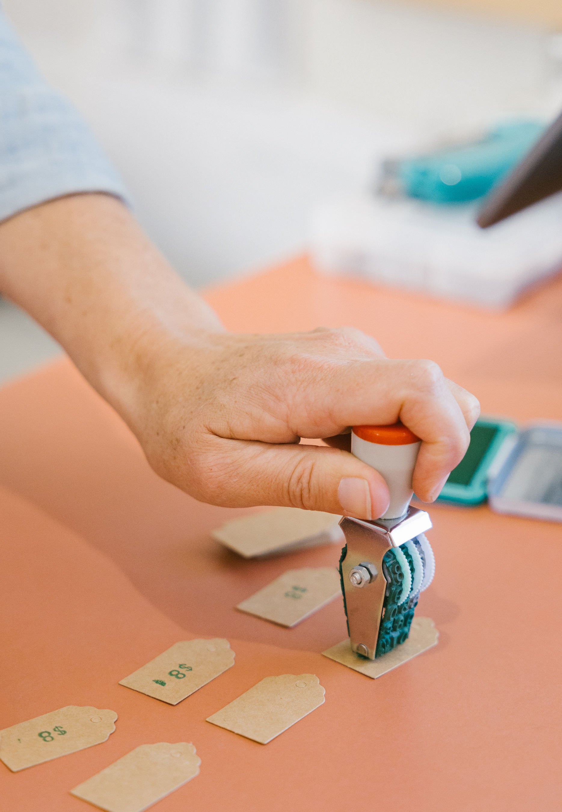 A person's hand using a price tag gun to attach a $8 price tag to a piece of paper on a pink surface with other similar tags nearby.