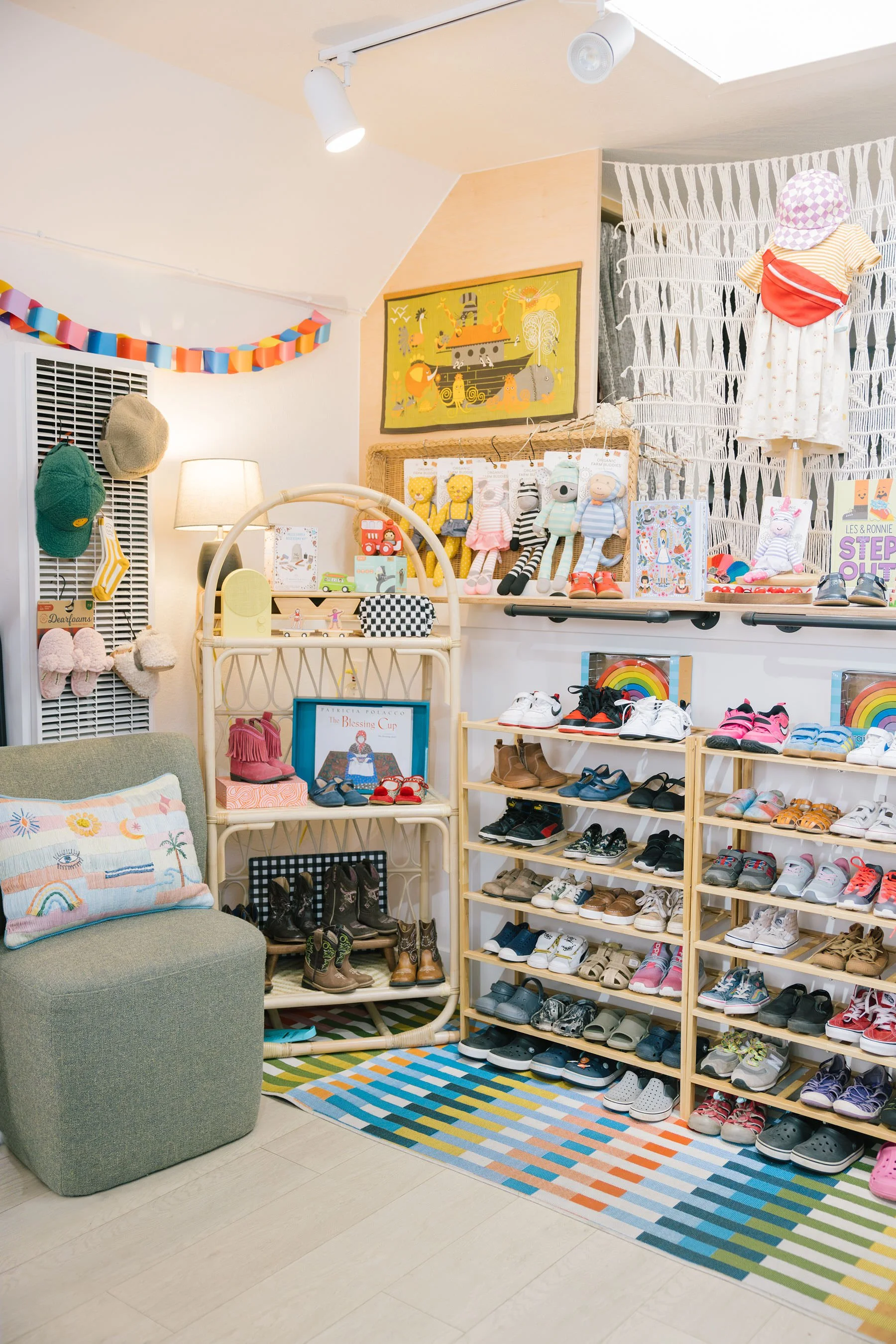 A children's shoe store with multiple wooden shoe shelves displaying sneakers, sandals, and boots. There is a green armchair with a colorful pillow, a multicolored striped rug, a white patterned wall, hats hanging on a grid, and a framed picture of a