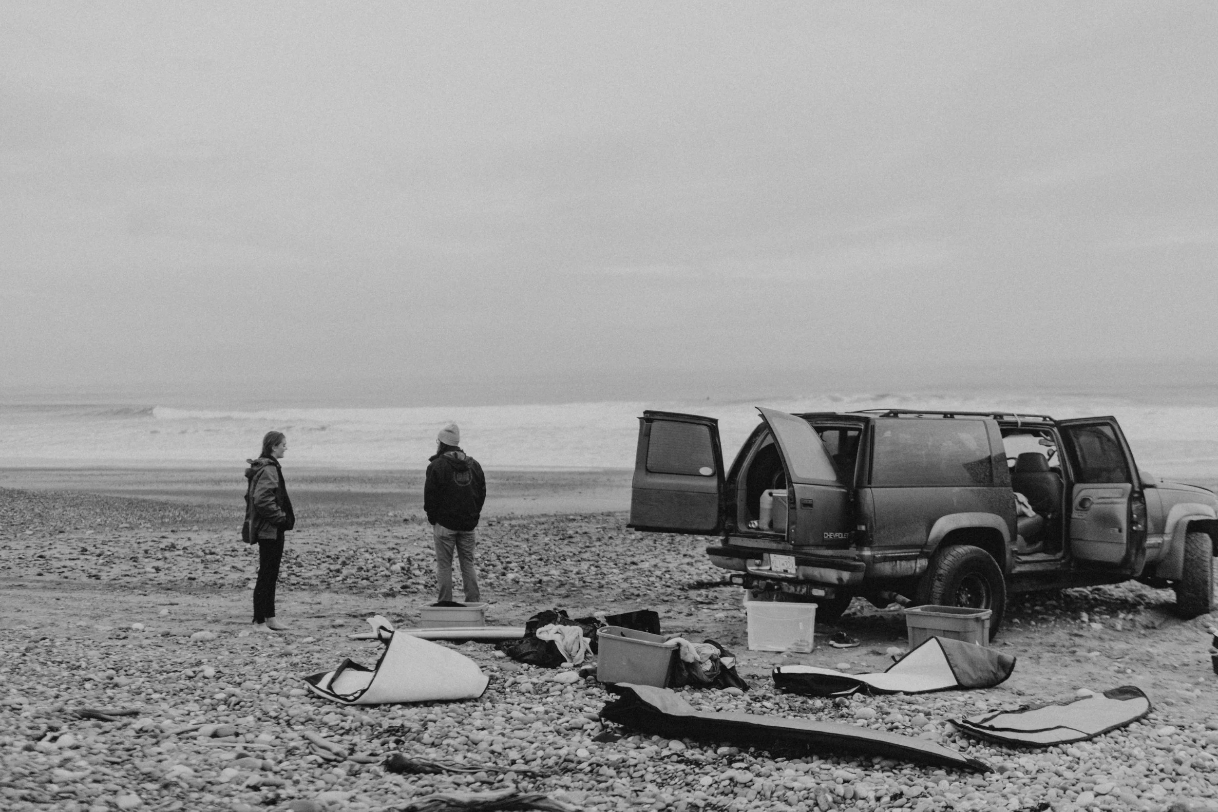 Haida Gwaii surf shop. Surfing Haida Gwaii. Surf Haida Gwaii. Surf rentals. Surfing. Surf beach Haida Gwaii. Three people standing on a pebble beach near a van,  overlooking an ocean or lake under a cloudy sky.