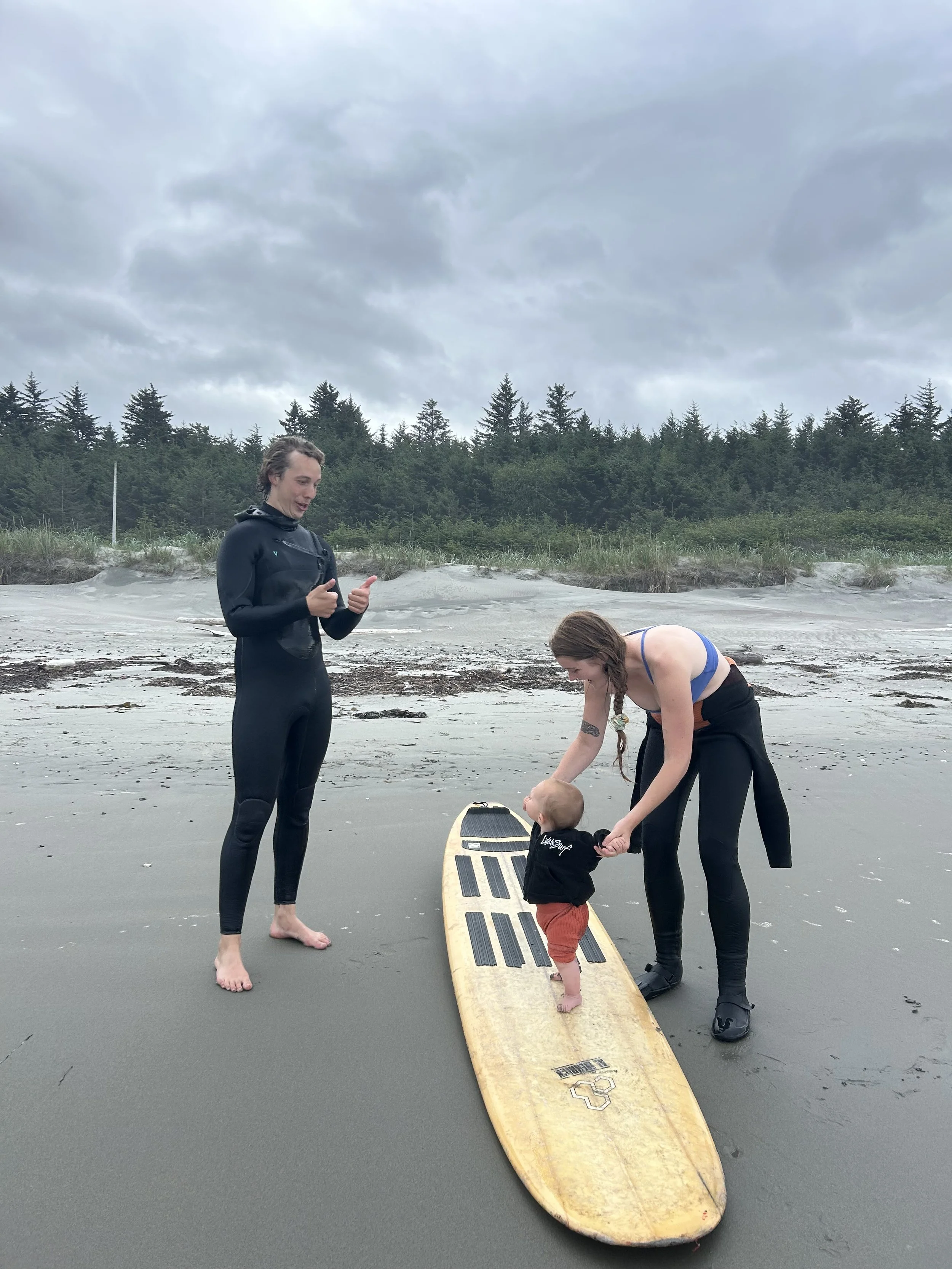 A woman in a black wetsuit and a woman with braids and black shoes helping a toddler stand on a yellow surfboard on a sandy beach with green trees in the background and cloudy sky overhead.