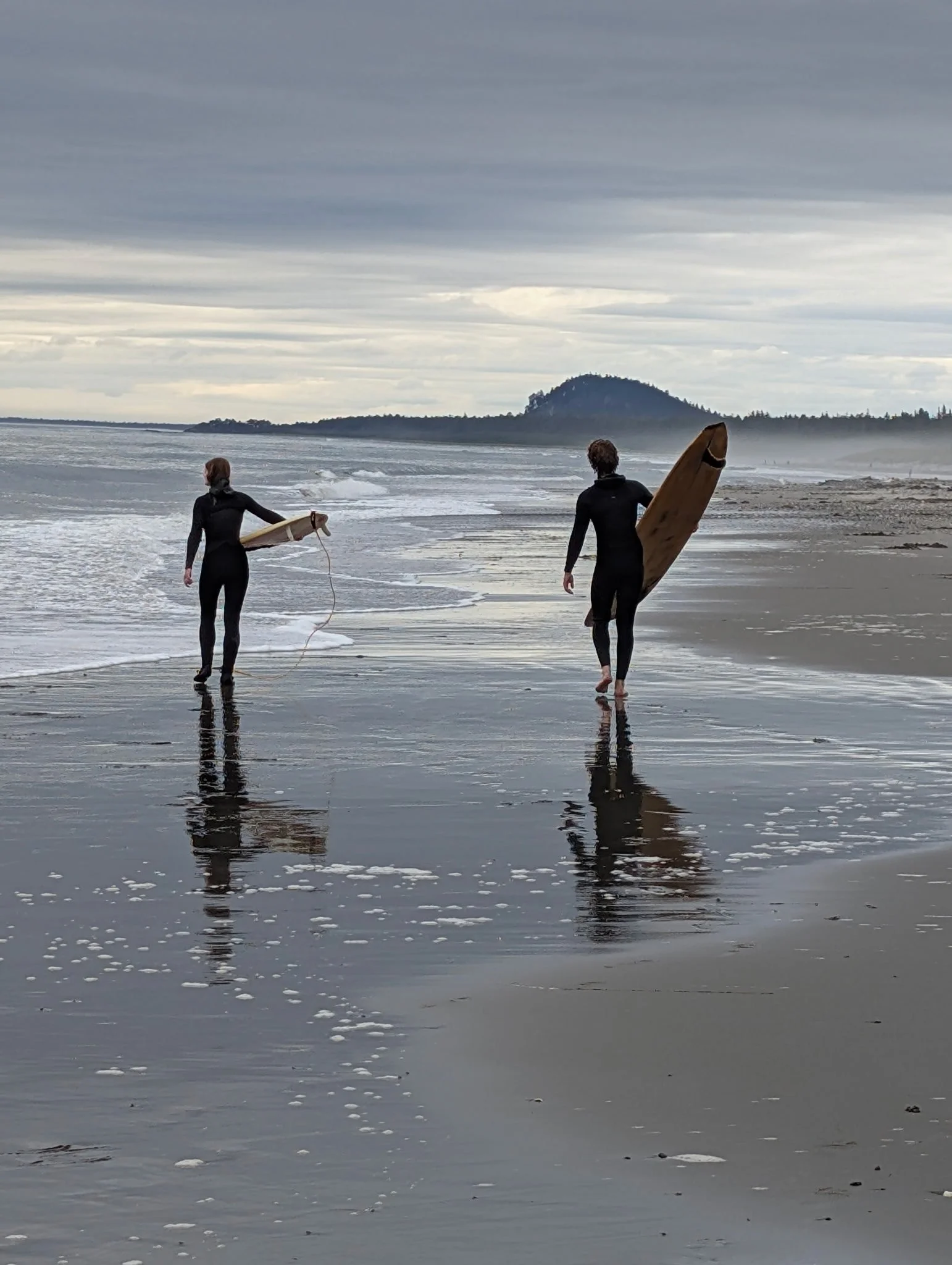 Two surfers in black wetsuits walking along the shoreline with surfboards under their arms, leaving footprints in the wet sand, with a cloudy sky and distant hills in the background.