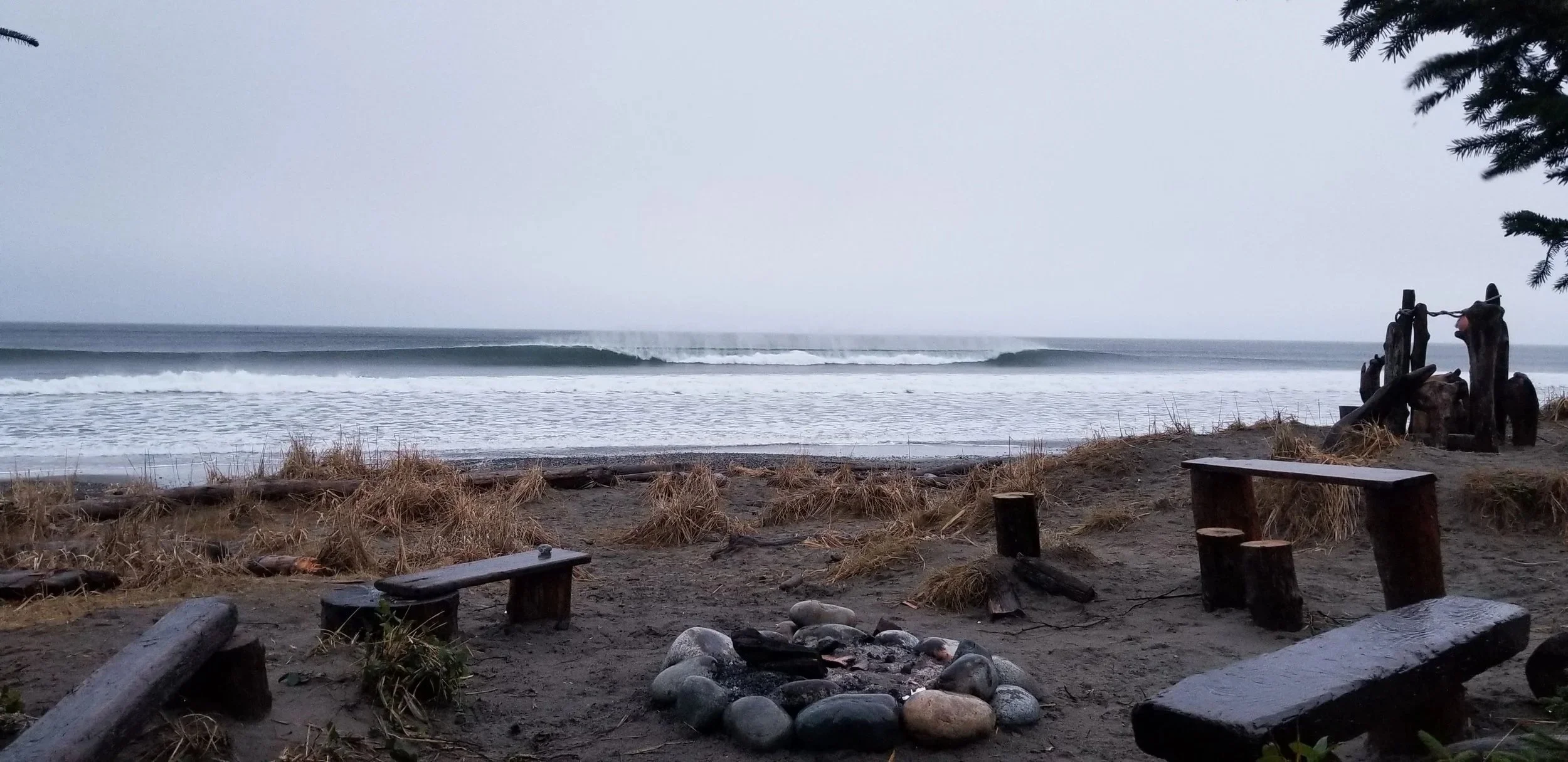A cloudy beach scene with a small bonfire circle and wooden benches in the foreground, tall grasses, and driftwood scattered on the sand, with ocean waves rolling in under an overcast sky. At Tagwaal on haida gwaii community surf spot
