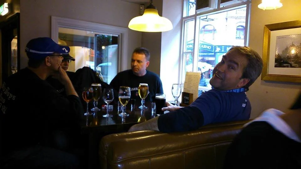 Vier Menschen sitzen in einer Bar oder einem Café, trinken Getränke, und unterhalten sich. Ein Mann im blauen Hemd sieht in die Kamera.